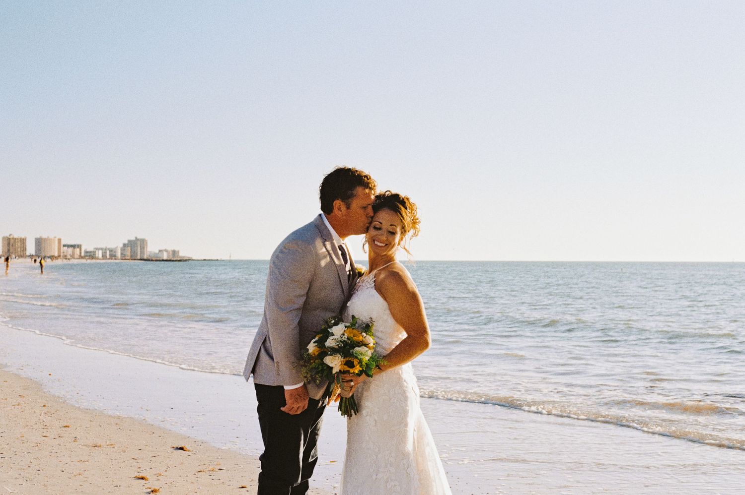 Groom in gray suit kissing bride holding sunflower bouquet on sunny beach at wedding.