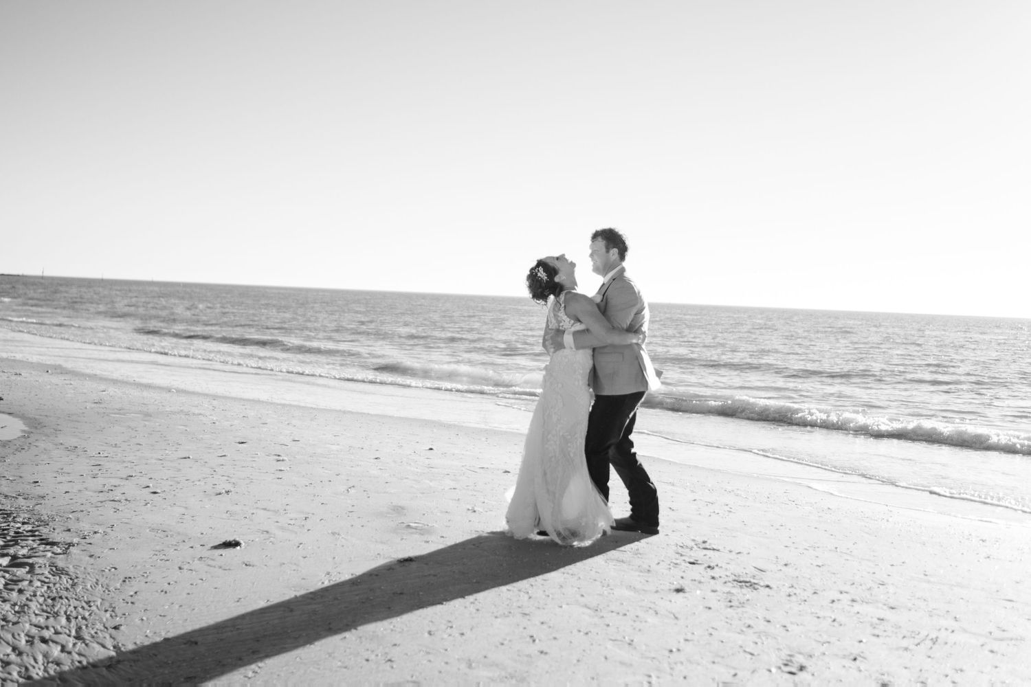 Black and white photo of bride and groom dancing romantically along the shoreline at the beach.