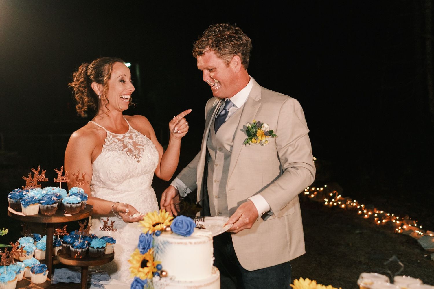 Bride and groom laugh joyfully while cutting their white wedding cake decorated with blue flowers at night.
