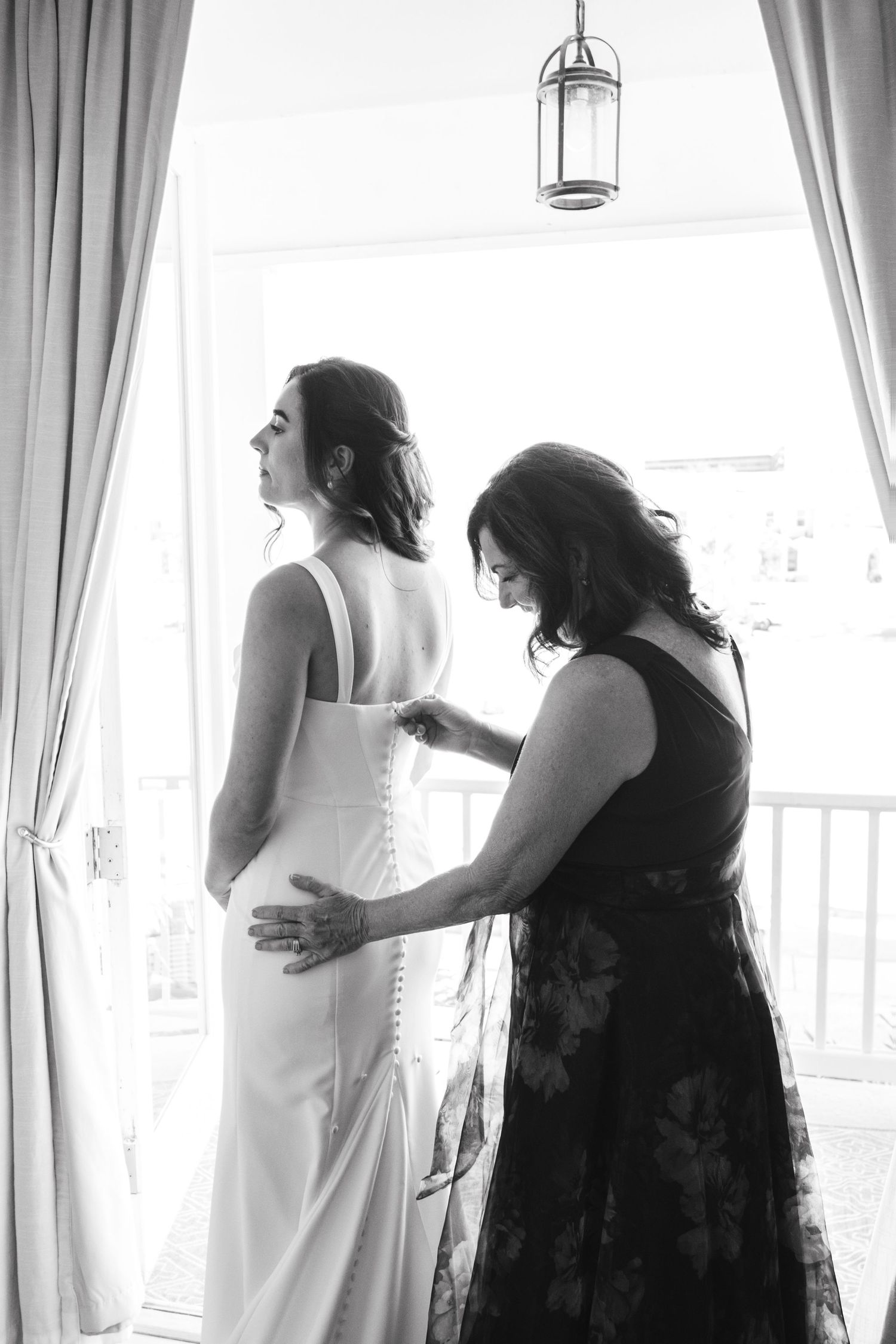 A tender moment as a woman in a black dress helps adjust a white wedding gown in front of draped windows.