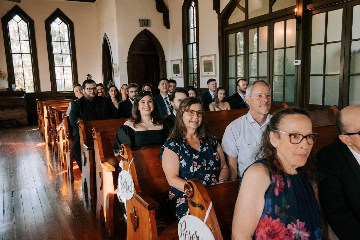 Wedding guests seated in wooden pews of a historic chapel during ceremony.
