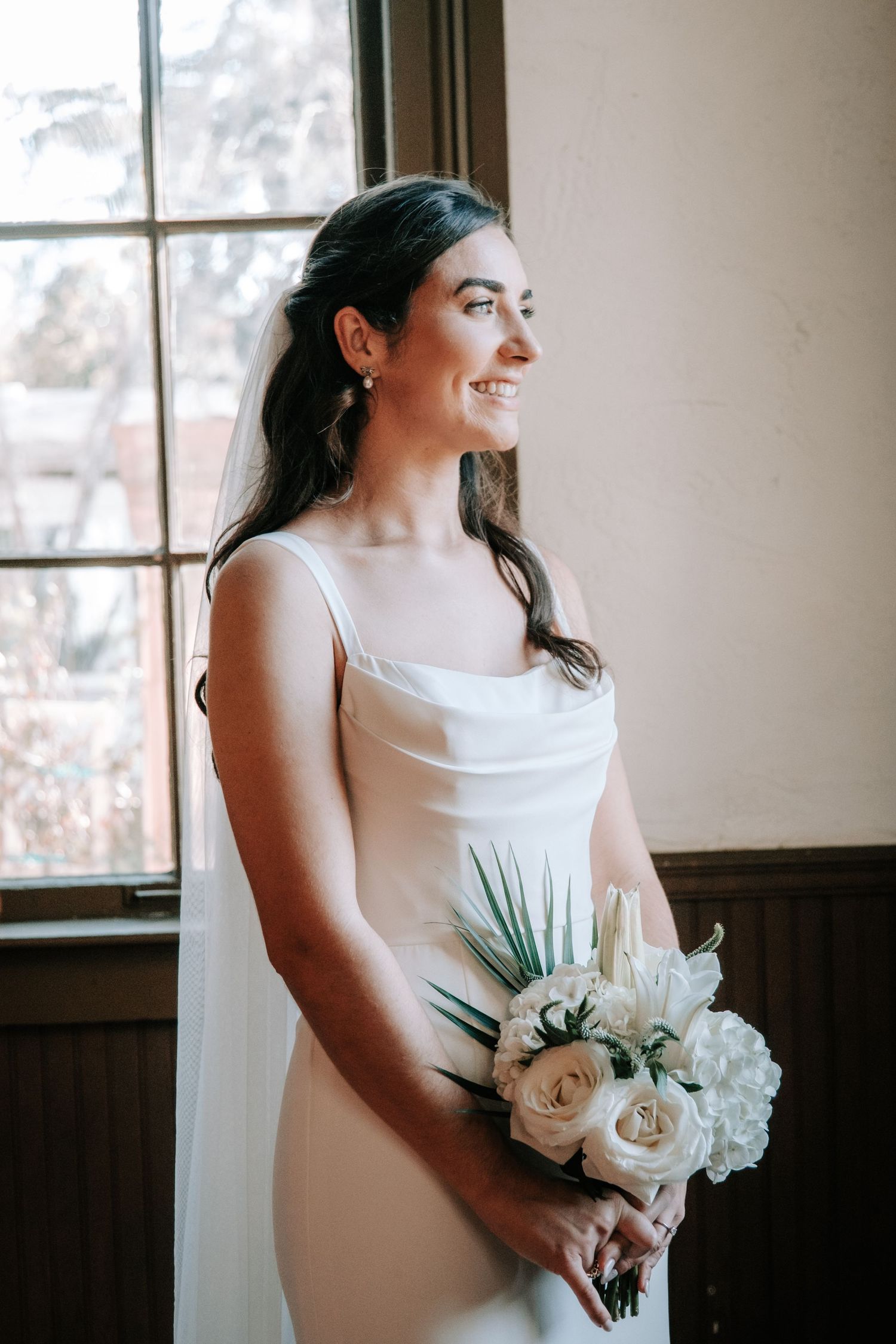 A bride in a sleek white dress holds a white floral bouquet while standing by a window.