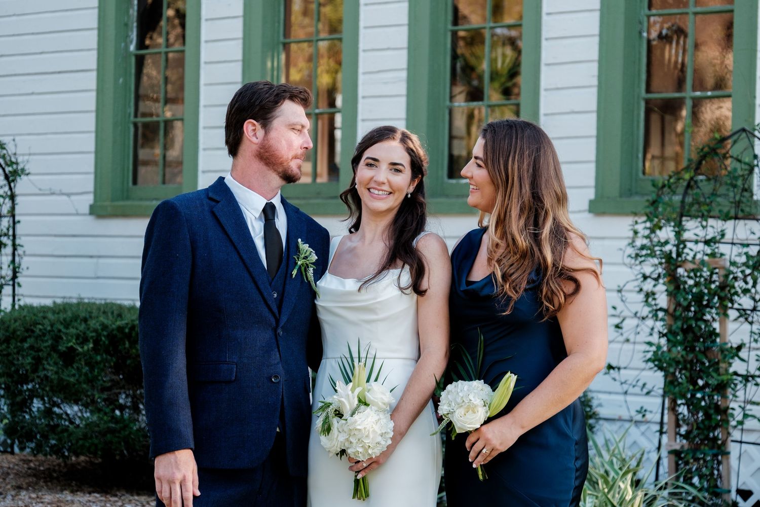 Wedding party poses outside historic white building with green trim and landscaping.