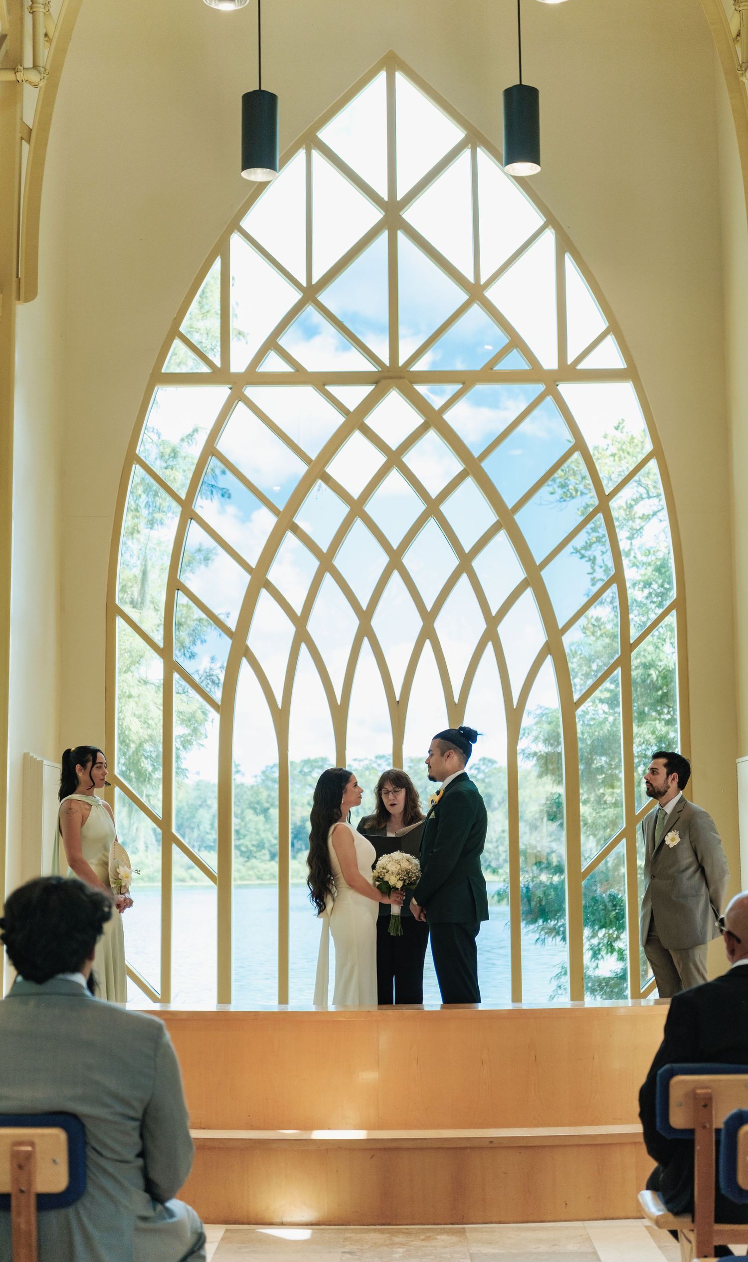 Wedding ceremony in progress inside chapel with dramatic geometric window and natural light.
