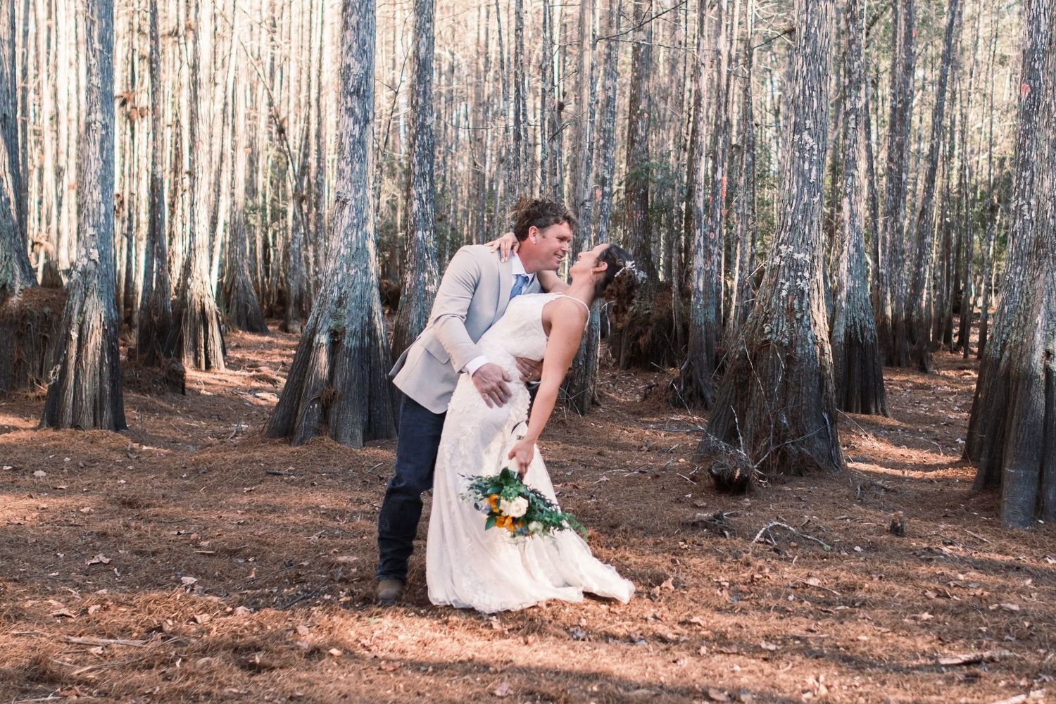 Bride and groom share a romantic dip kiss among tall trees in a forest wedding photo.