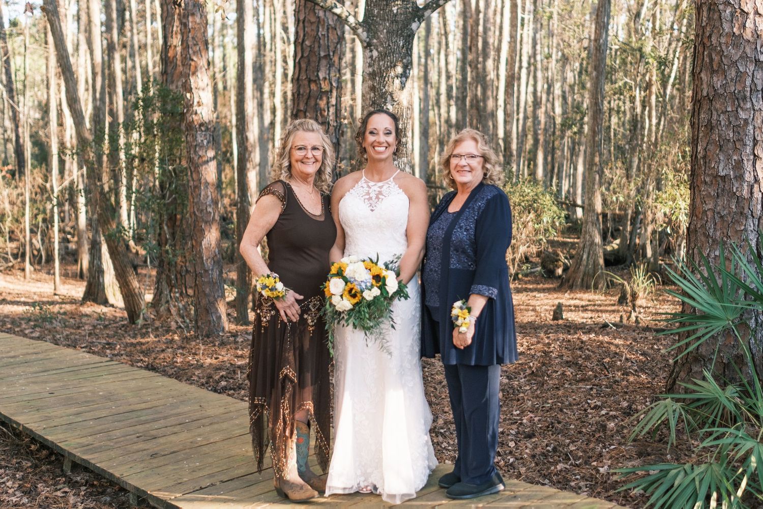 Bride in lace halter gown holds sunflower bouquet between two smiling women on a forest boardwalk.