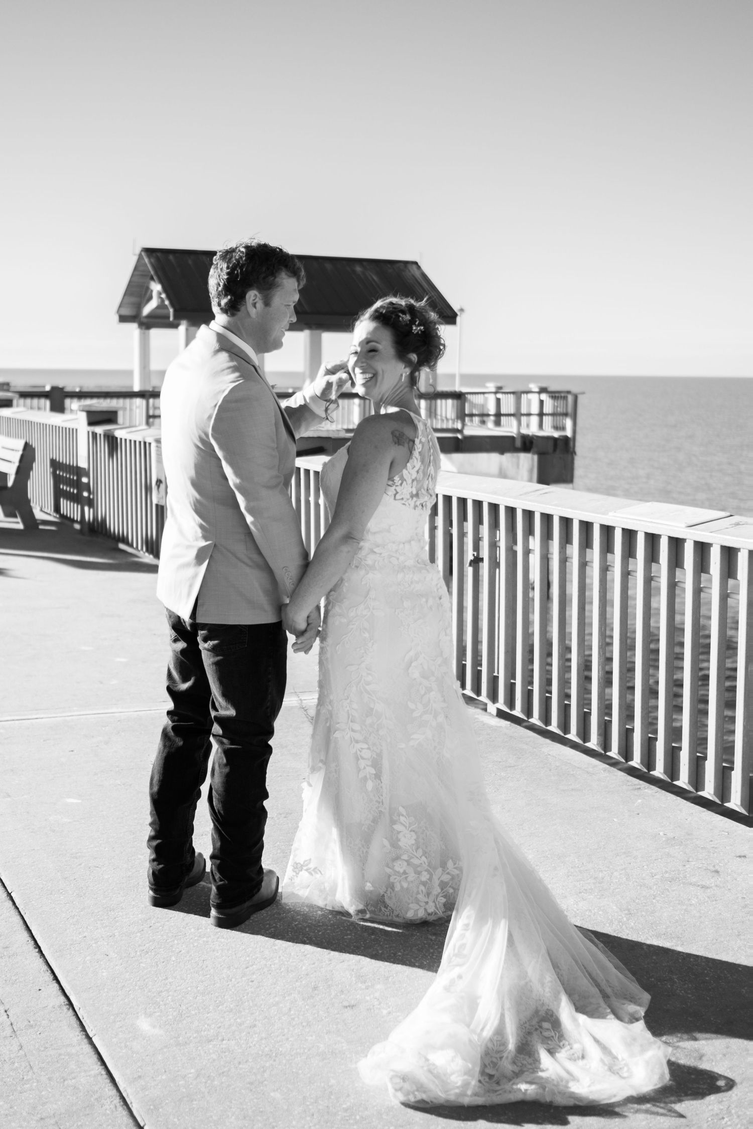 Black and white photo of bride and groom embracing on waterfront pier on wedding day.