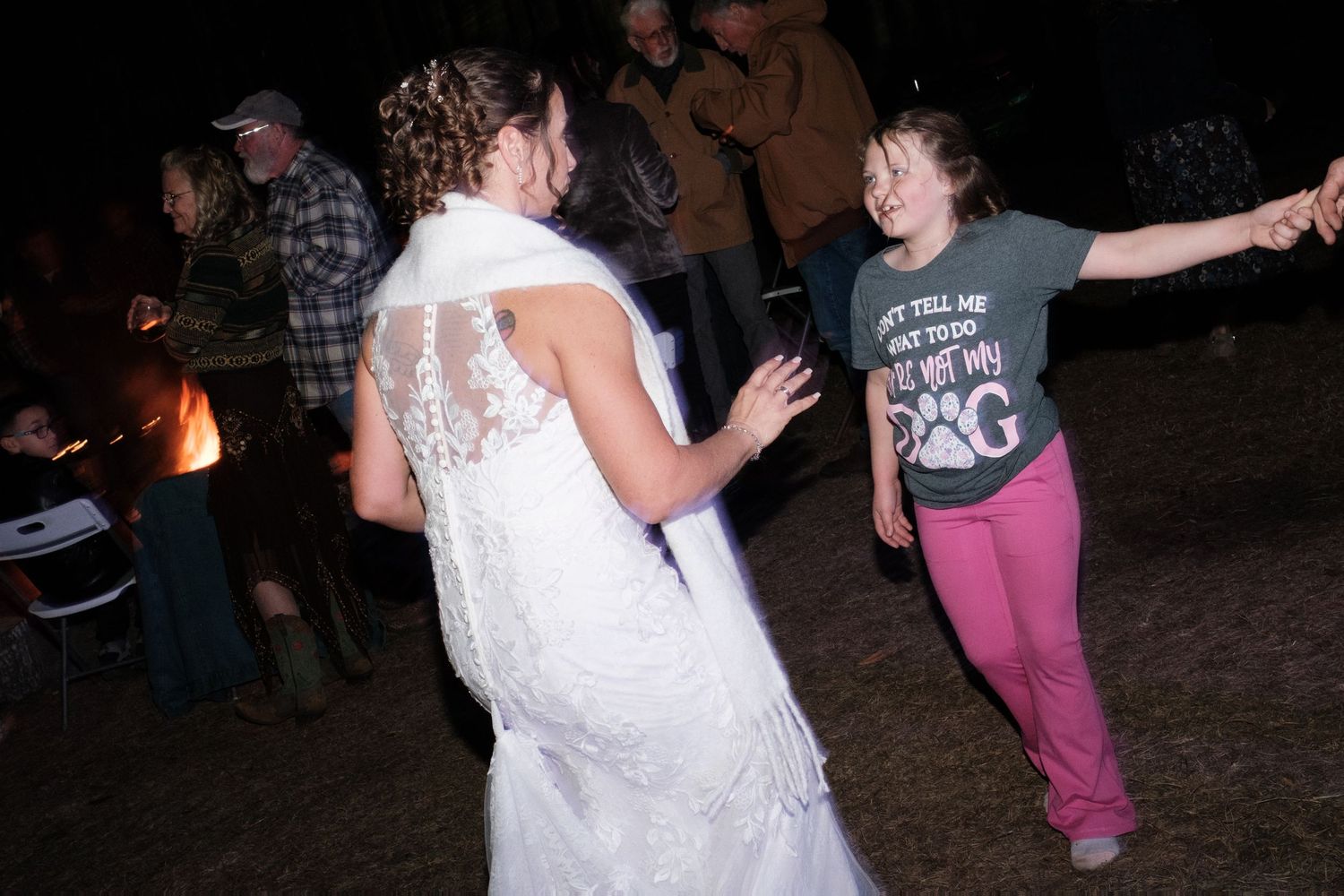 Bride in a lace wedding gown dances energetically with a young girl at an outdoor evening wedding reception.