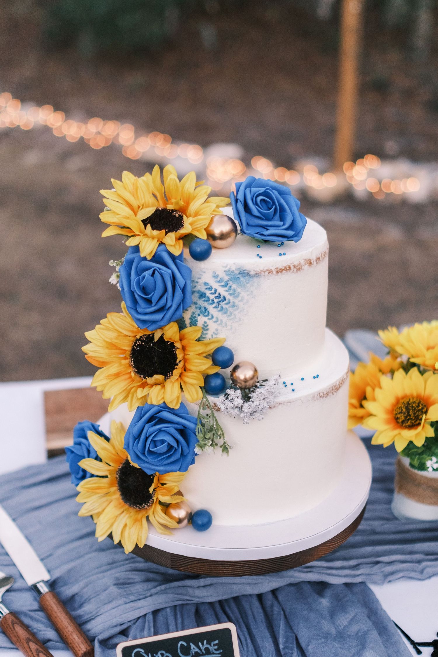 Elegant two-tier white wedding cake decorated with blue roses, sunflowers, and gold accents on a draped table.