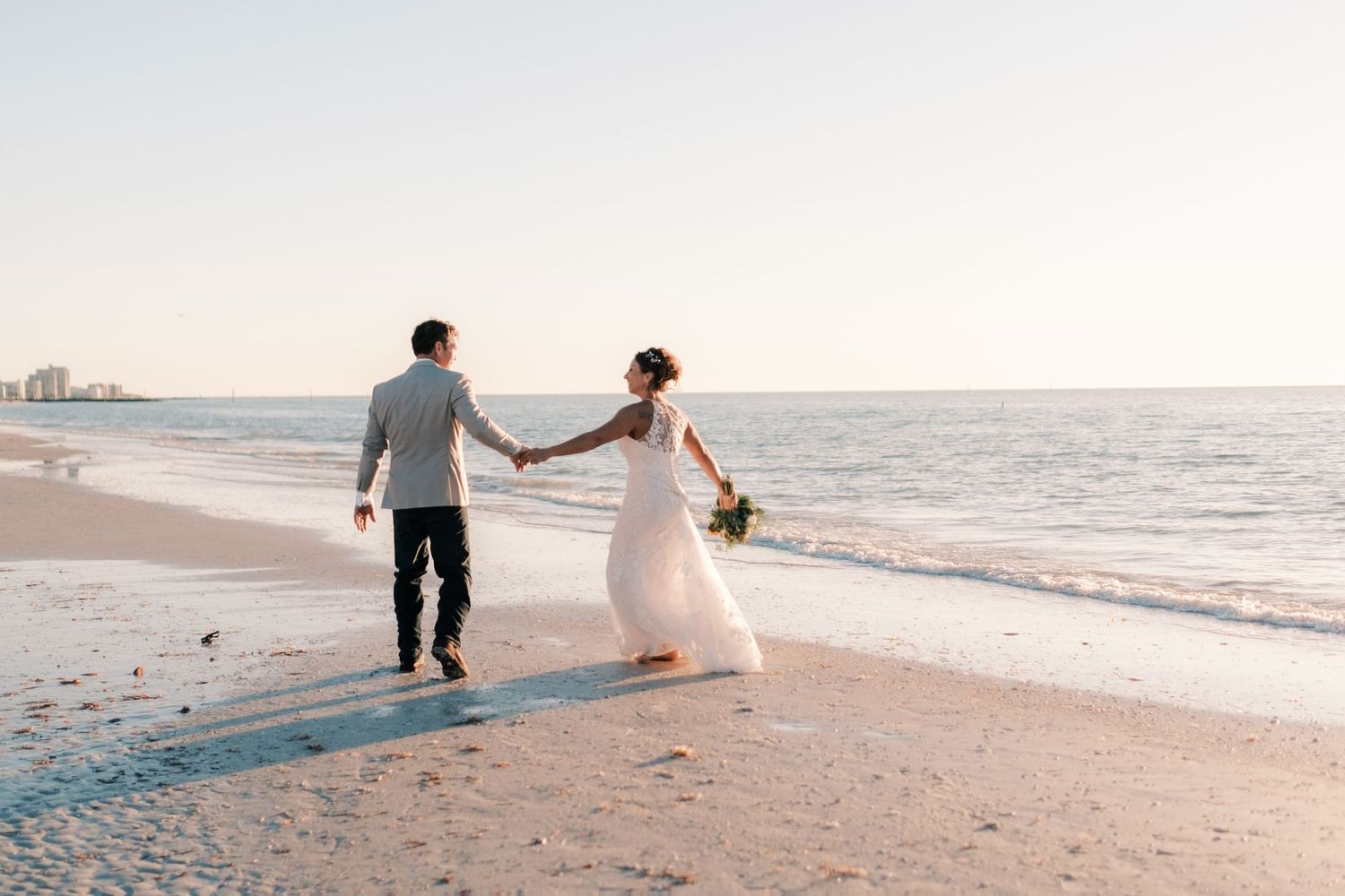 Bride and groom walk hand in hand along a serene beach shoreline at sunset on their wedding day.