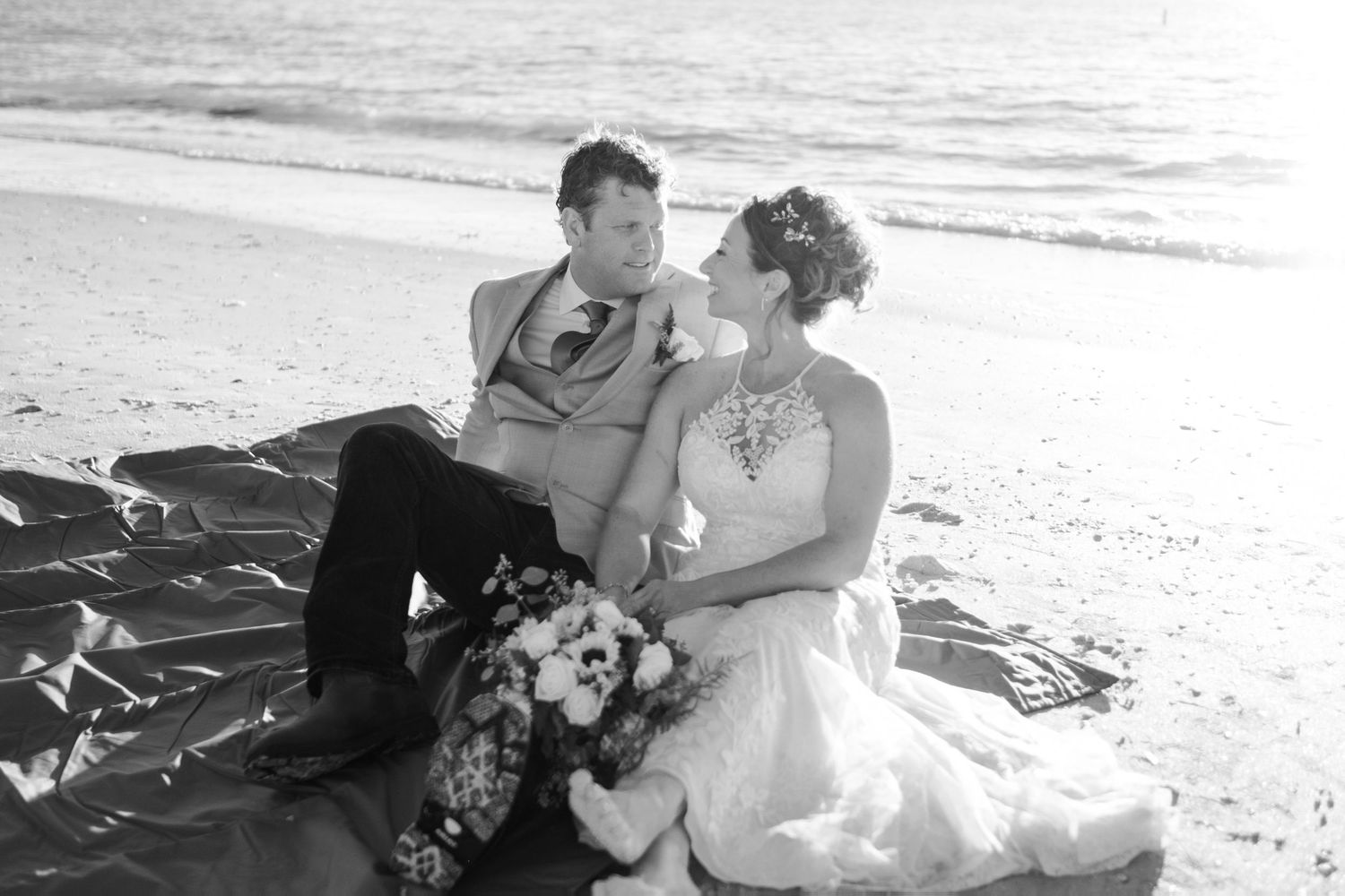 Black and white photo of bride and groom sitting together on a beach blanket, sharing a romantic moment.