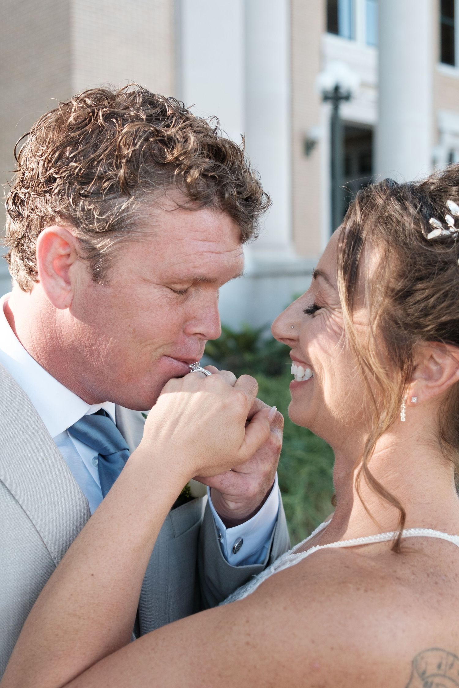 Groom kisses bride's hand tenderly as she laughs joyfully during their wedding day portraits.