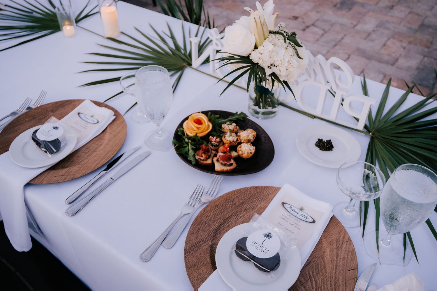 An elegant table setting with palm leaves, white flowers, and wooden chargers for a wedding reception.