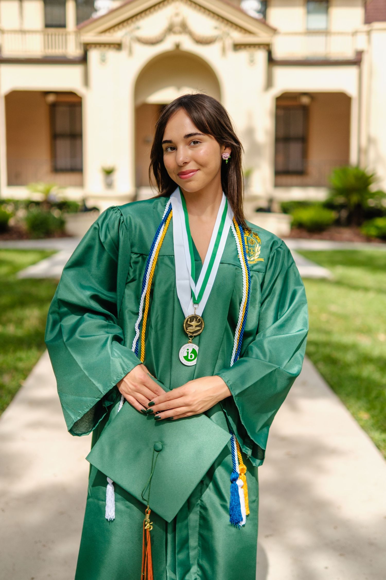 Graduate in emerald green commencement gown and honor cords poses with medals in front of ornate building entrance.