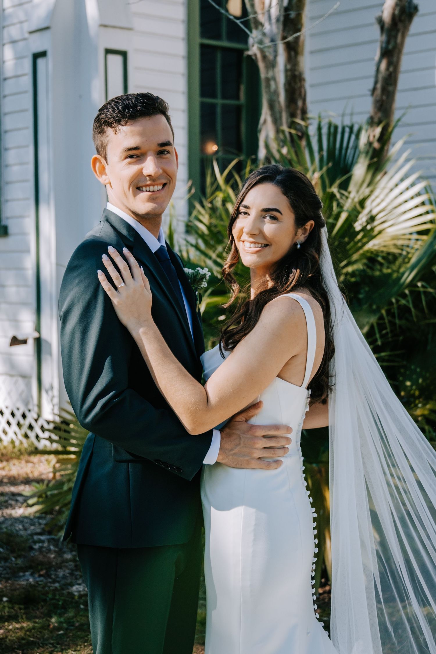 Bride in white dress and groom in dark suit pose together outside a white building.