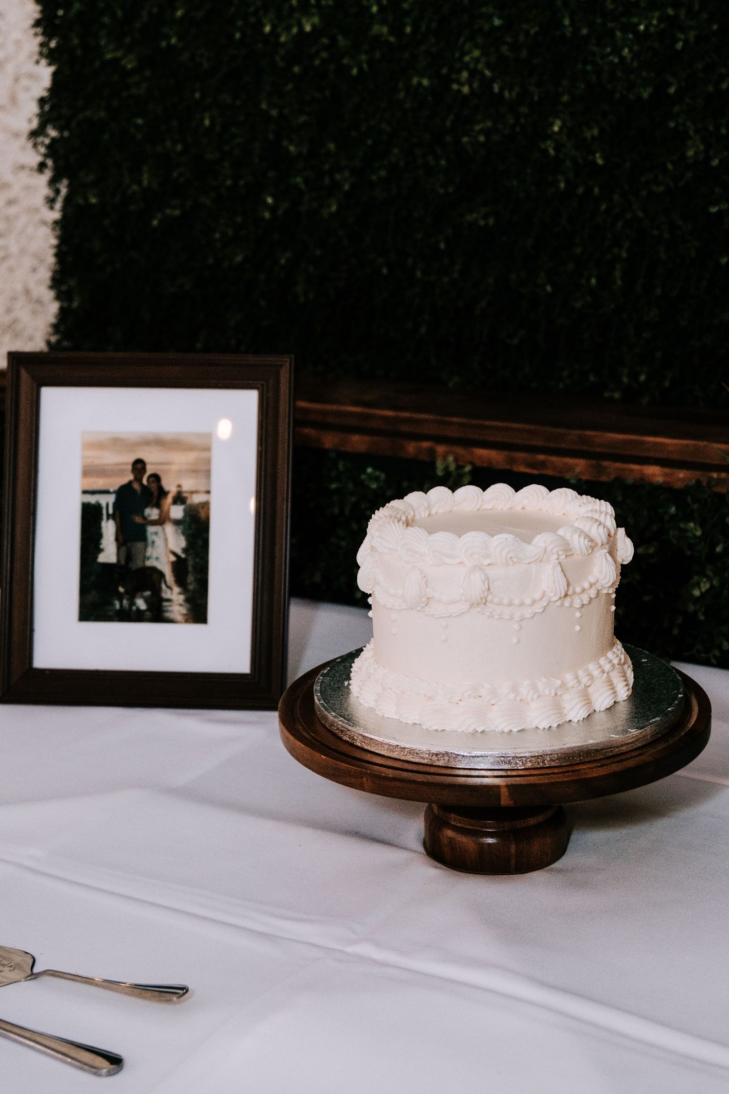 A simple white wedding cake displayed on a wooden stand against a dark background.