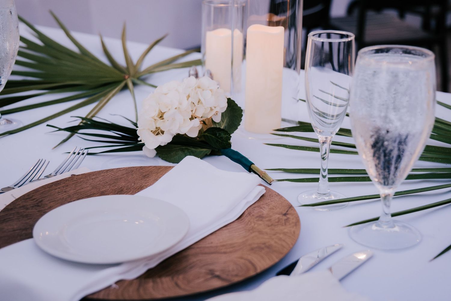 Wedding reception table details with palm leaves and place settings on white linens.