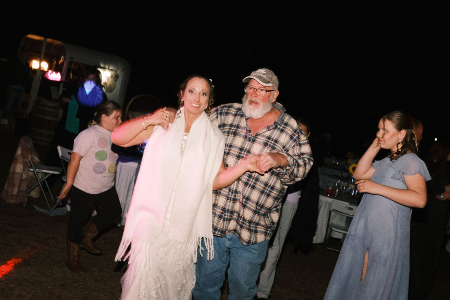 Bride in white dress and shawl dances with a guest in plaid shirt at an outdoor evening reception.
