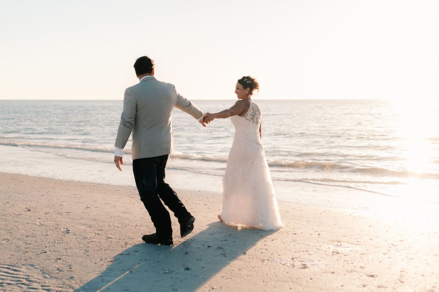 Newlywed couple walks hand in hand along a serene beach at sunset in wedding attire.