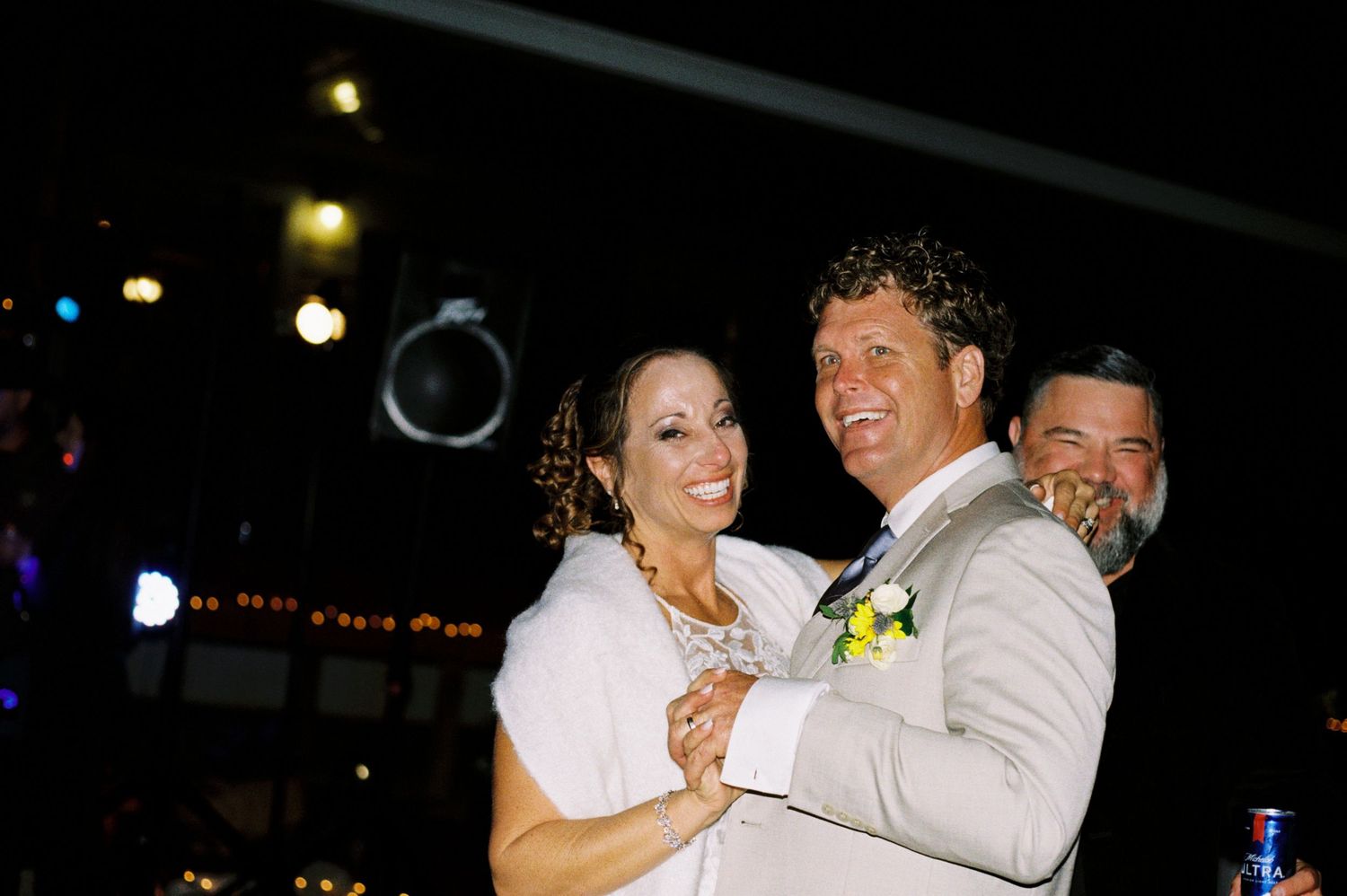 Smiling bride and groom share first dance under string lights at evening wedding reception.
