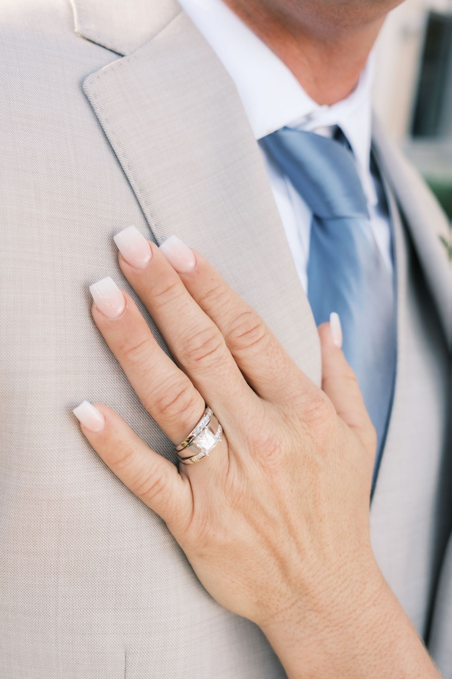 Bride's hand with wedding ring rests on groom's tan suit jacket with blue tie.
