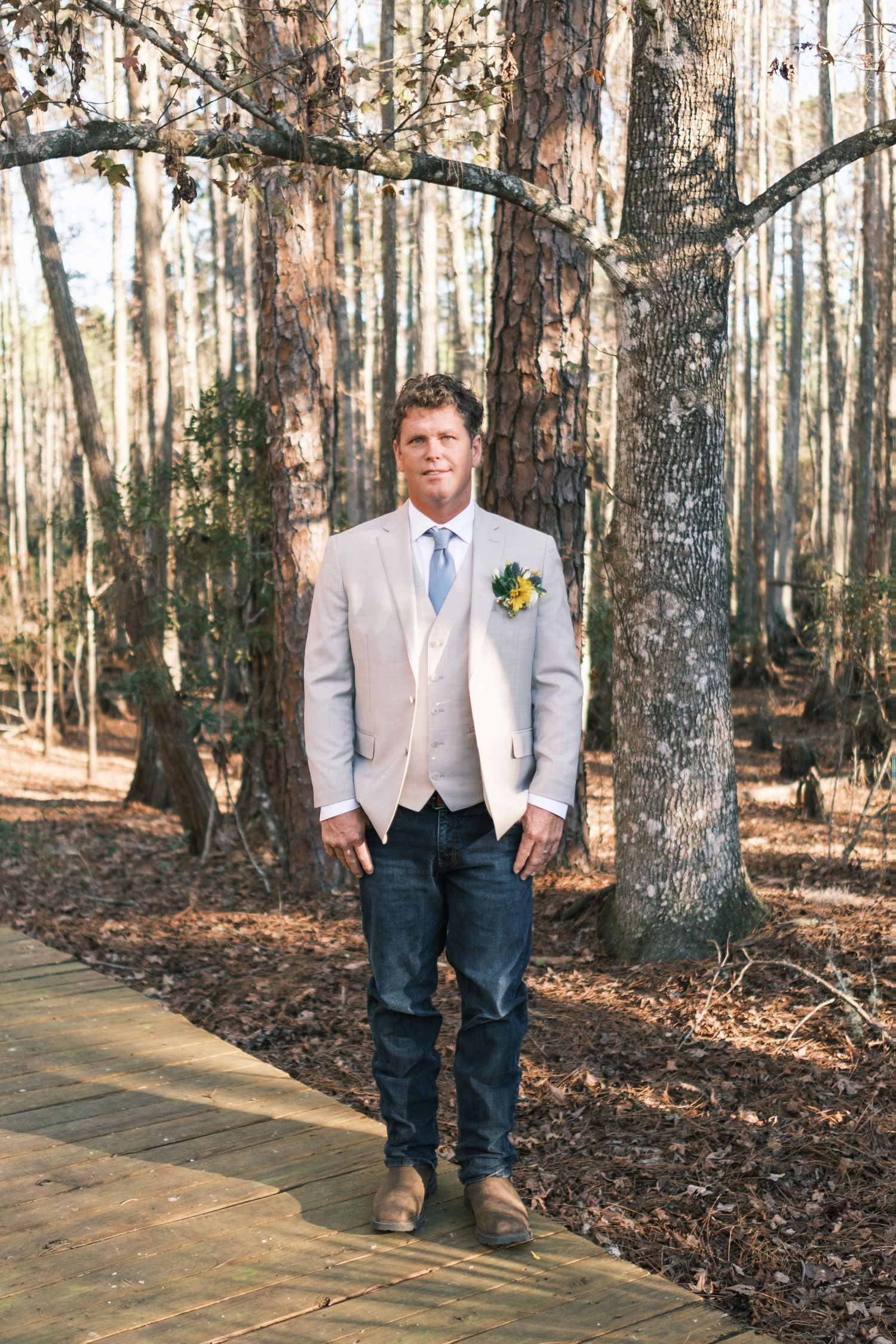 Dapper groom in a tan suit, blue tie, and jeans stands confidently on a wooden path in a sunlit forest.