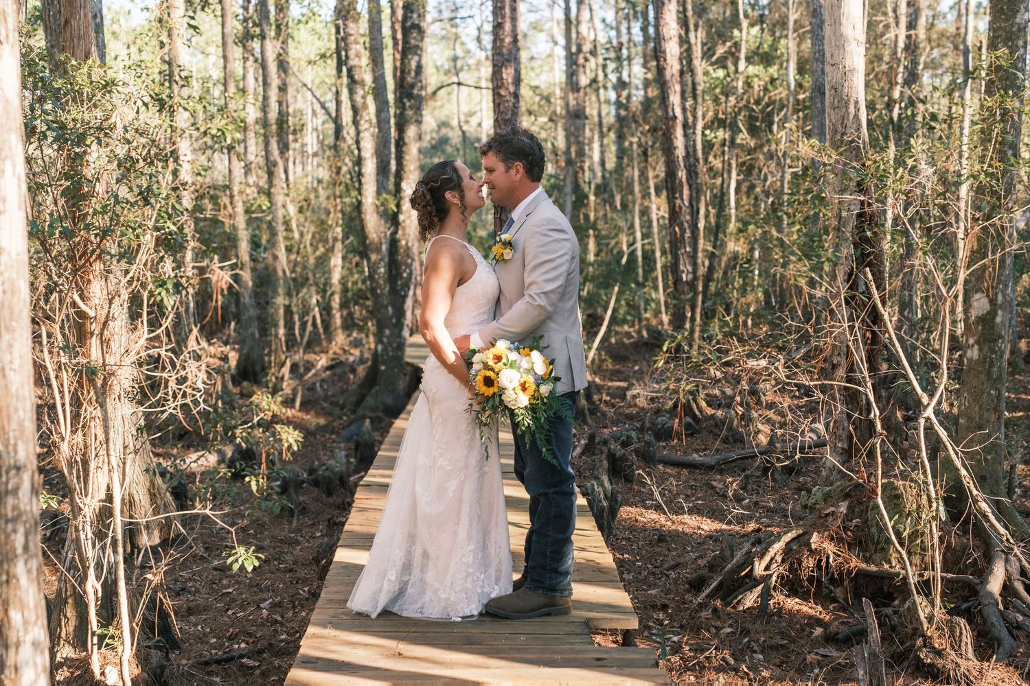 Bride and groom share an intimate moment on a wooden bridge surrounded by a sunlit forest.