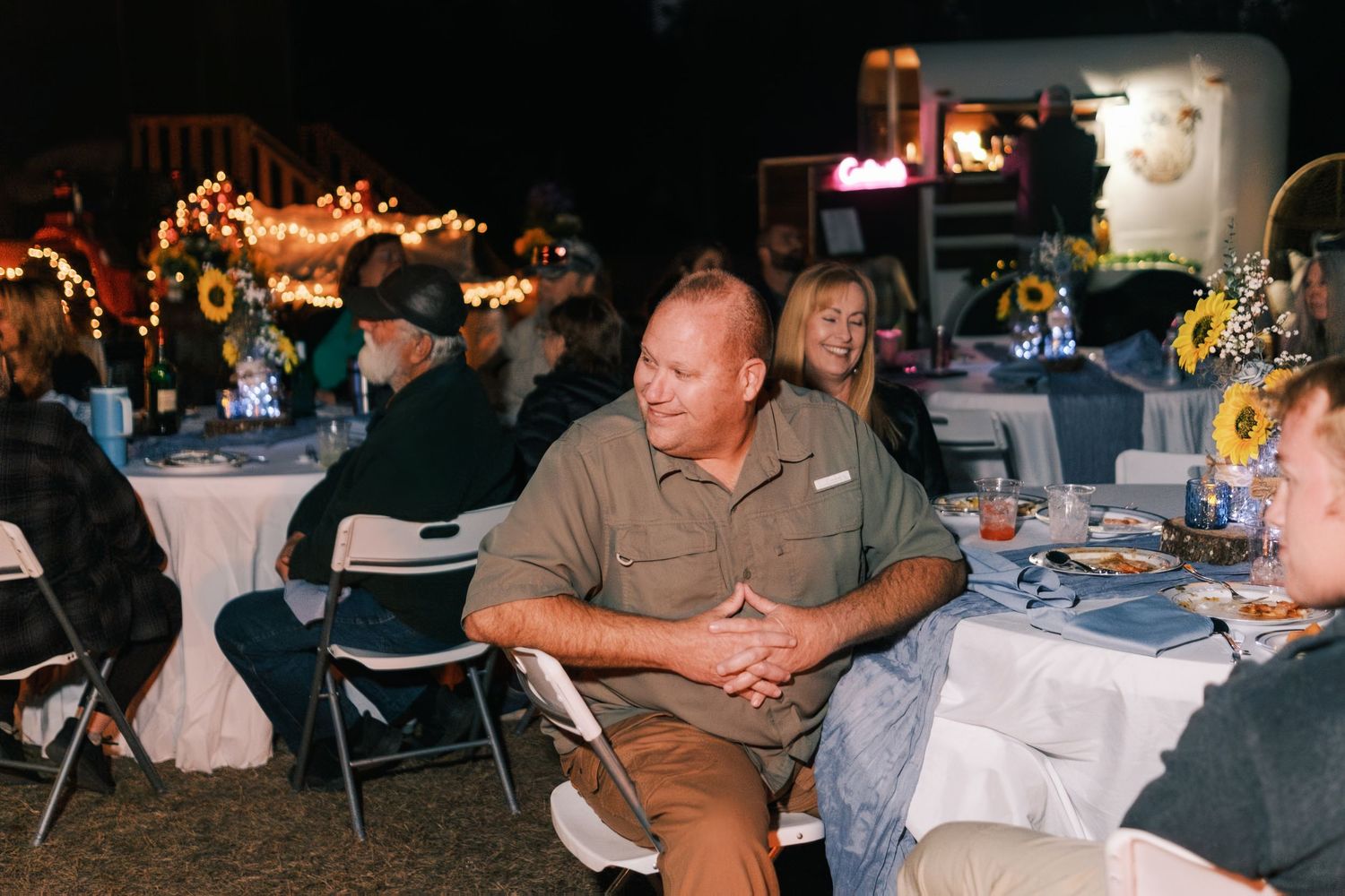 Wedding reception guests seated at decorated tables with sunflower centerpieces under warm string lights.
