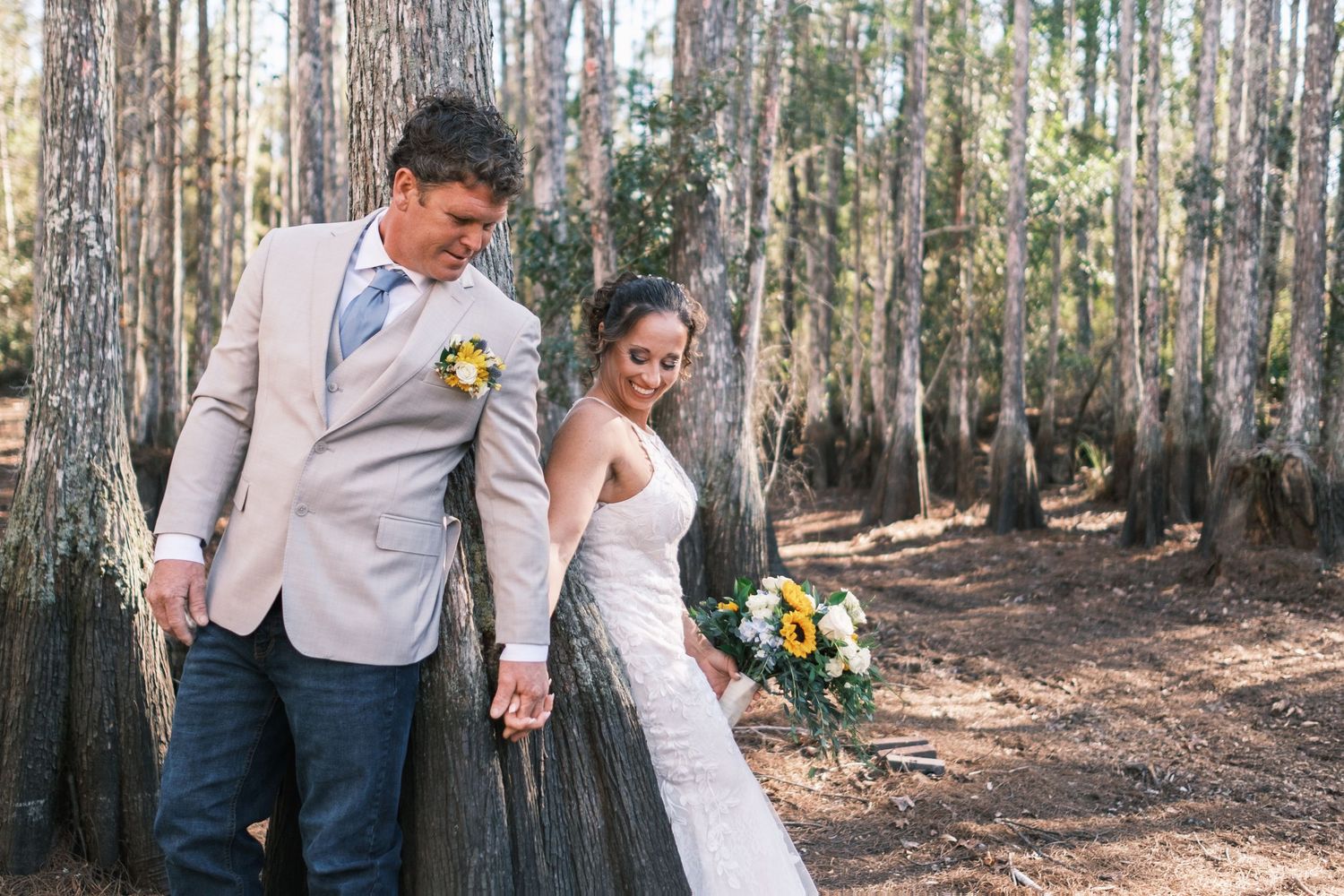 Bride and groom holding hands walking through a pine forest, bride holding yellow and white bouquet.