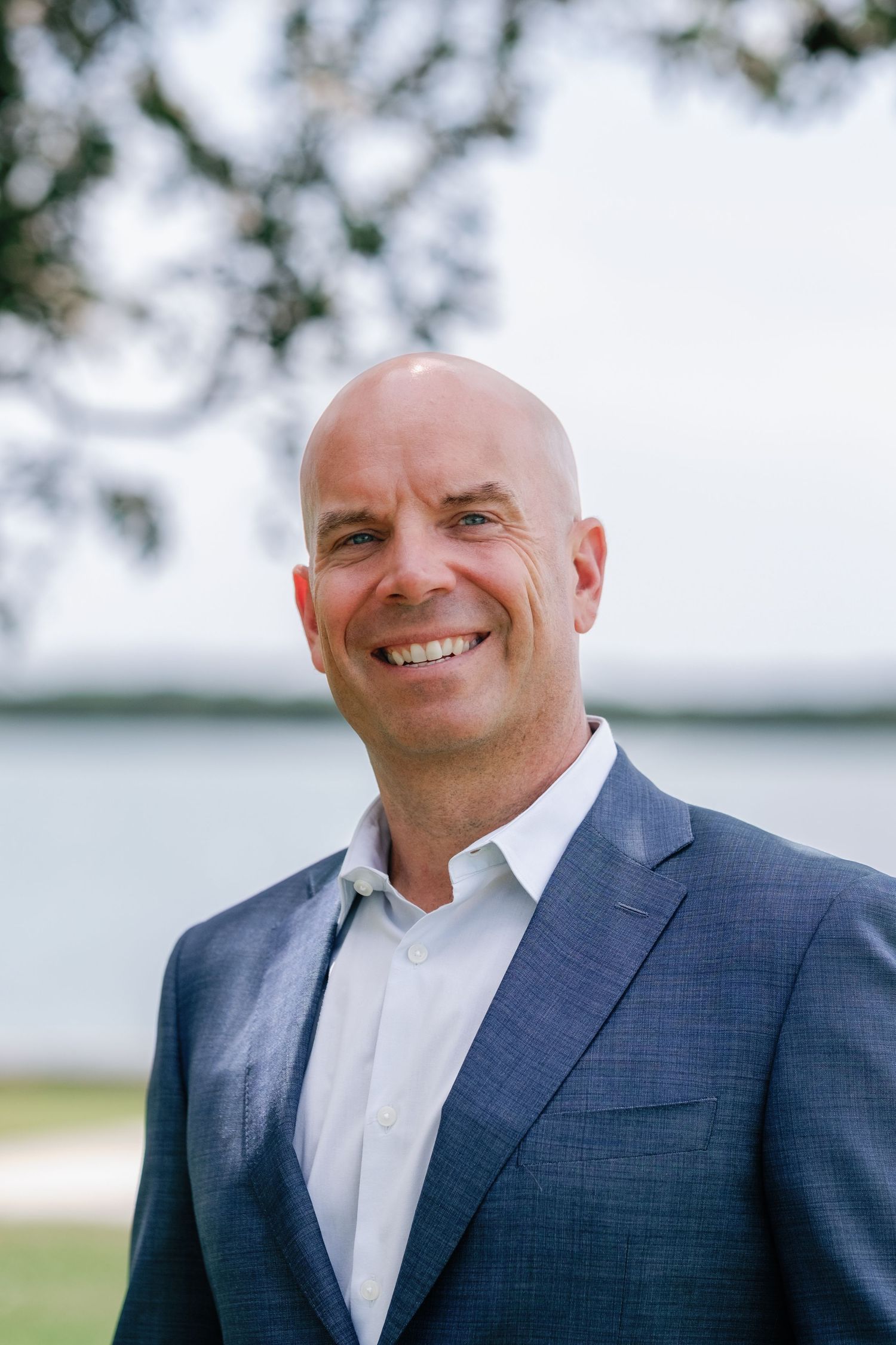 Smiling bald man in blue blazer and white shirt posing outdoors near a lake with trees in background.