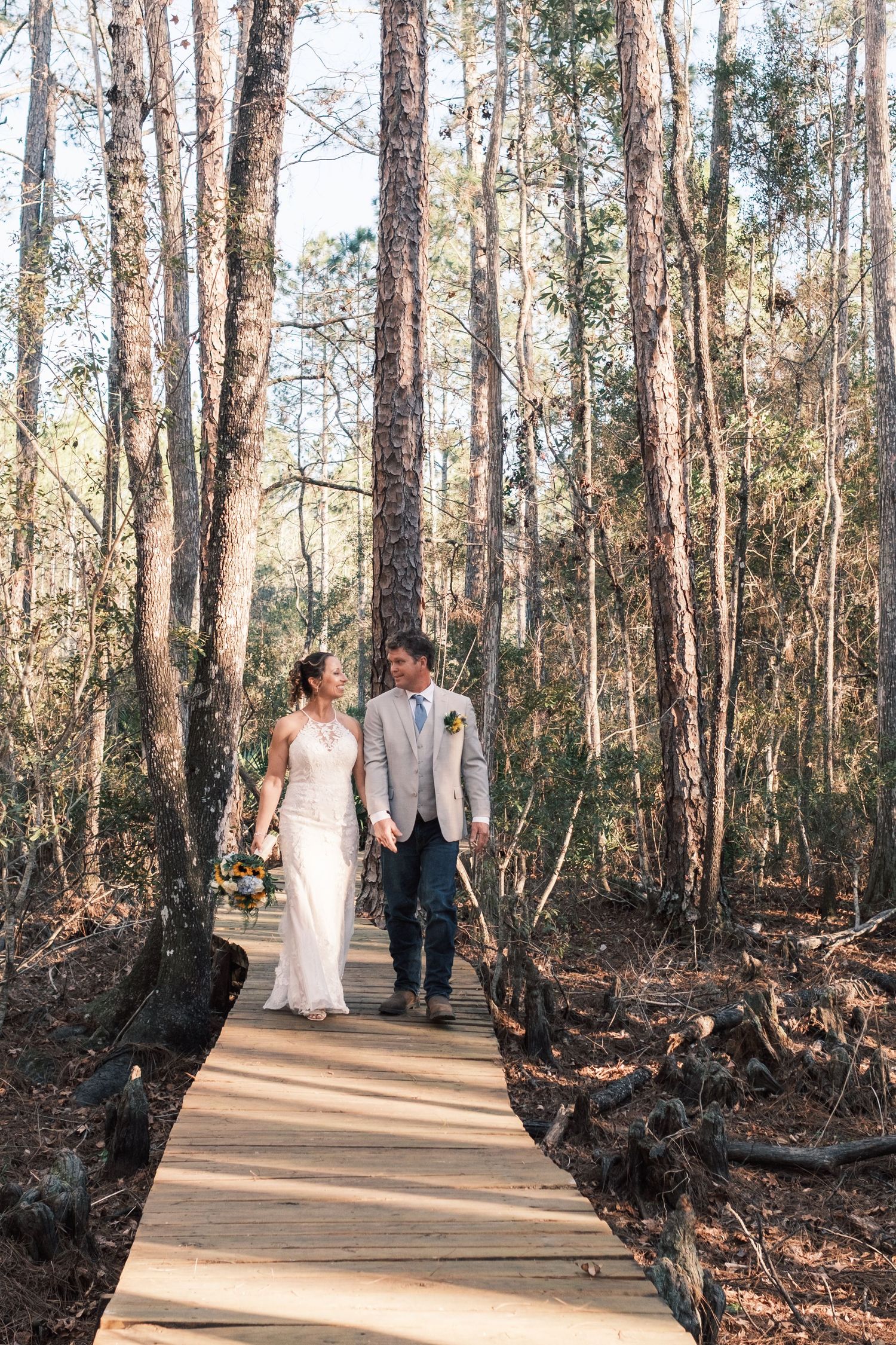Bride and groom walking hand in hand on a wooden boardwalk through a sunlit forest on their wedding day.