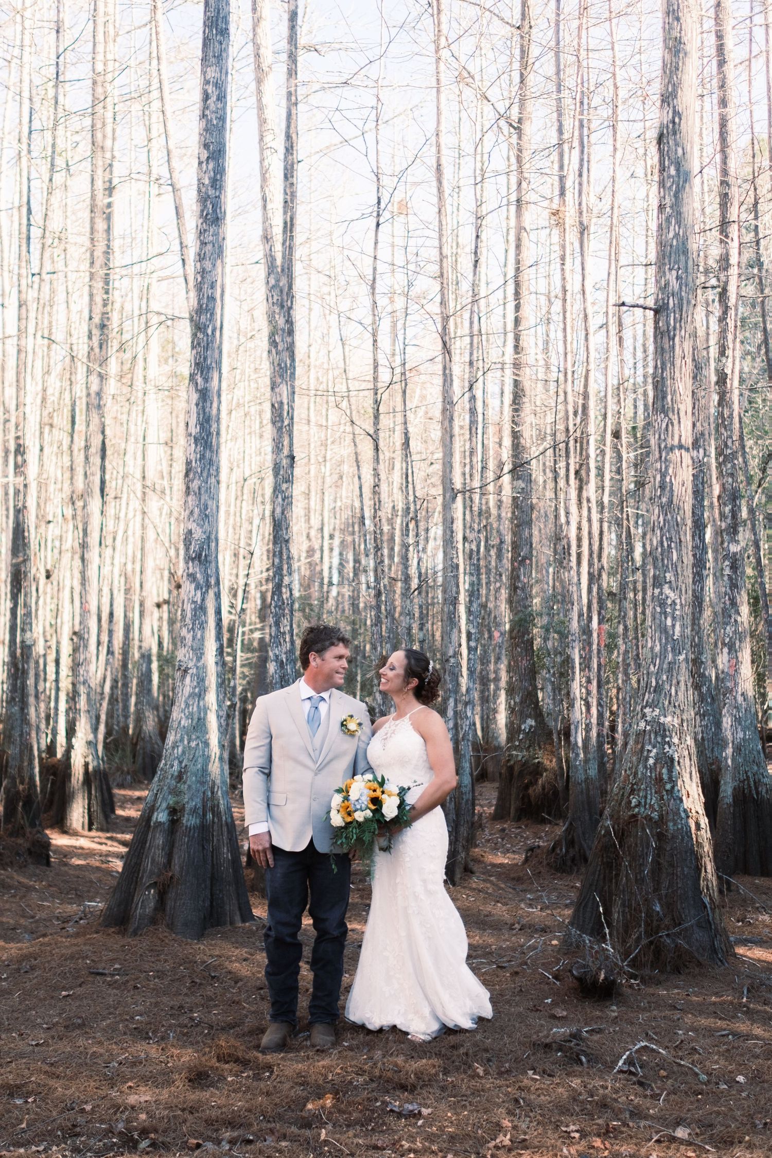 Bride and groom stand together among tall bare trees in a forest, bride holding a colorful bouquet.