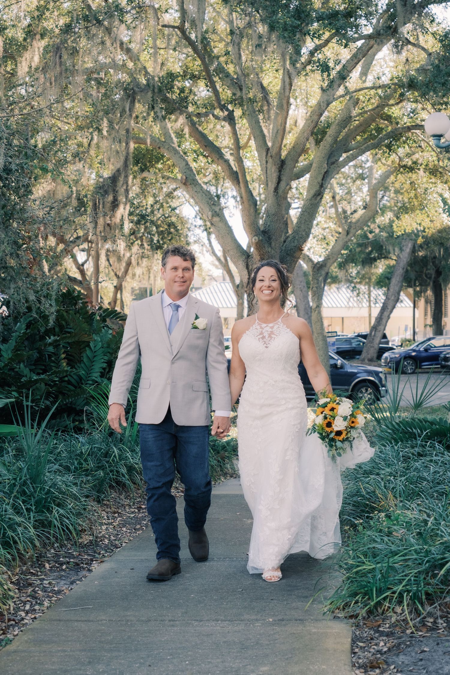 Bride and groom walking hand in hand through tree-lined garden path.