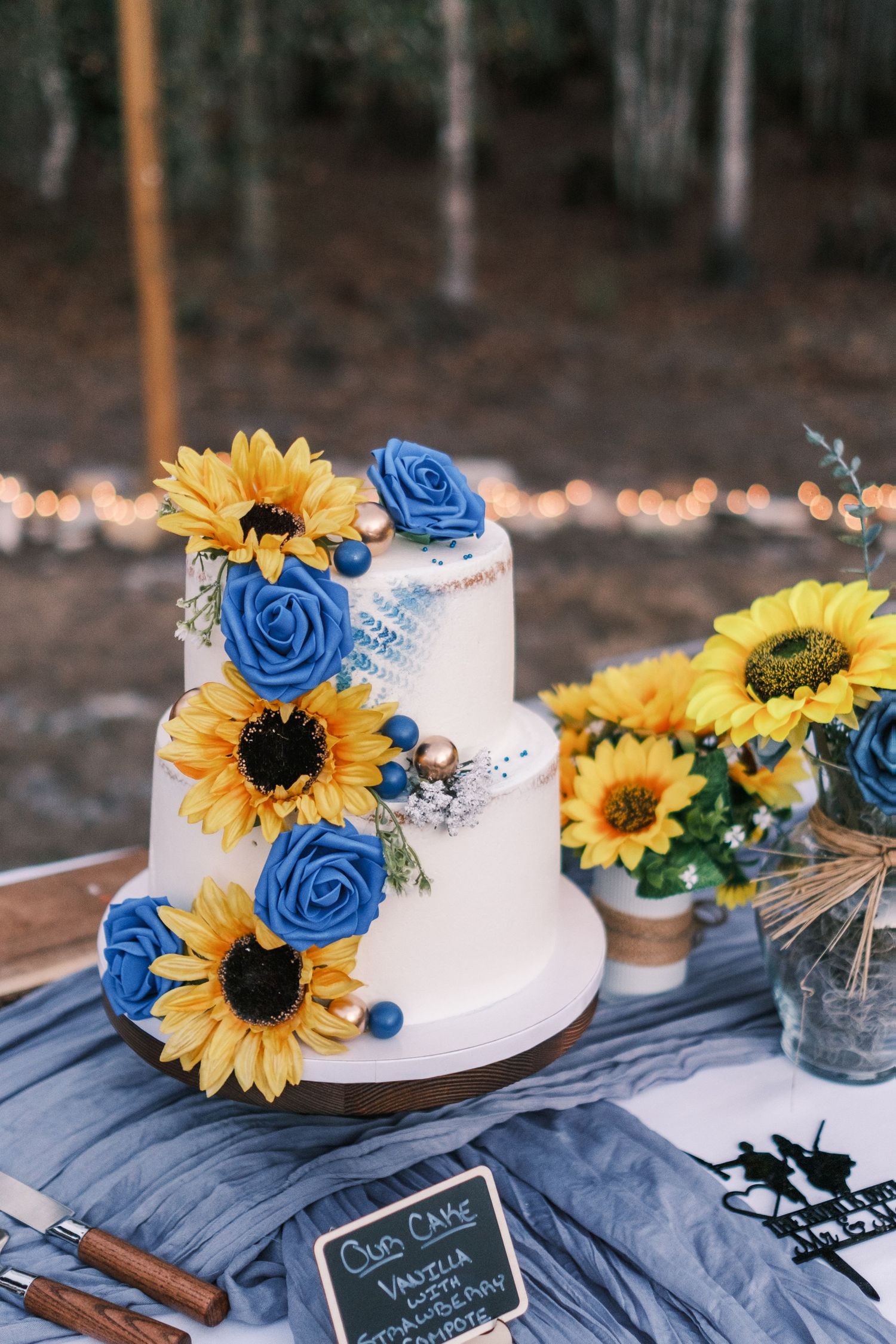 Two-tier white wedding cake decorated with sunflowers and blue roses on a wooden cake stand.