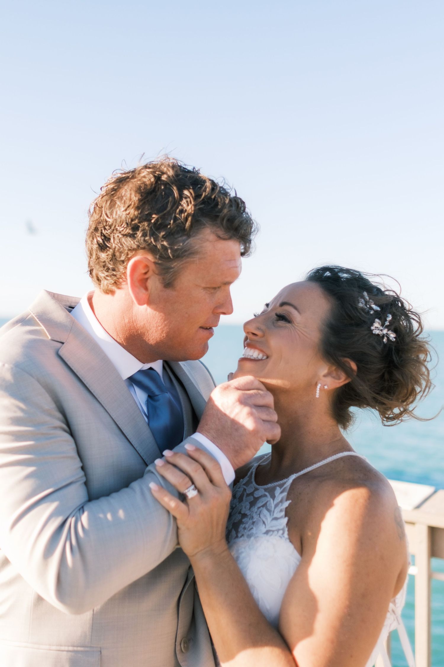 Groom tenderly holds bride's face as she smiles joyfully during romantic outdoor wedding portrait by the ocean.