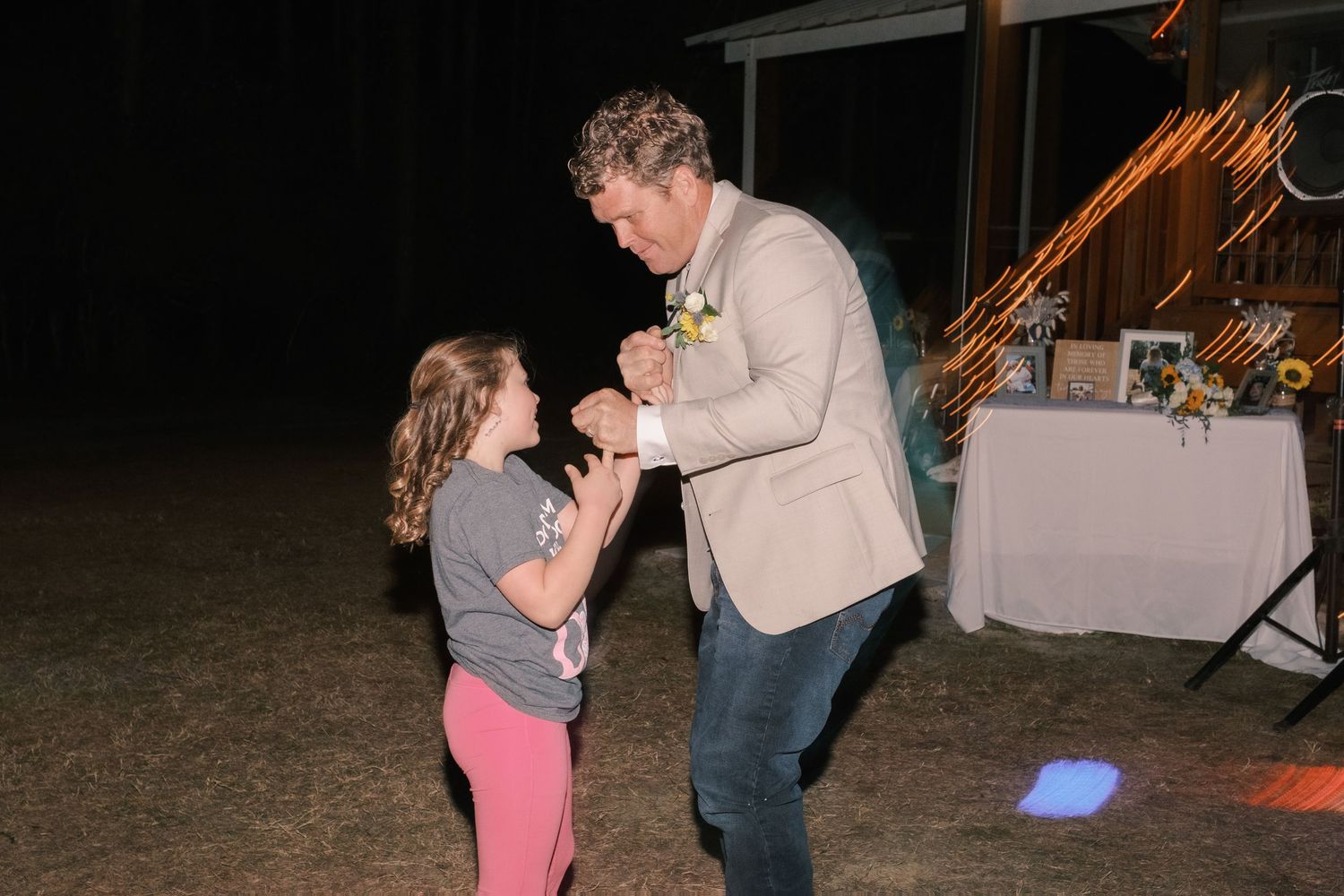 Groom shares a sweet dance with a young girl under colorful lights at wedding reception.