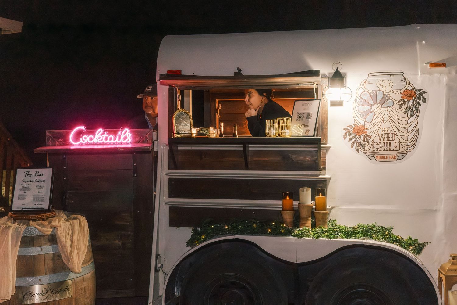 Vintage white trailer converted into a cocktail bar with neon sign glowing at an outdoor wedding reception.