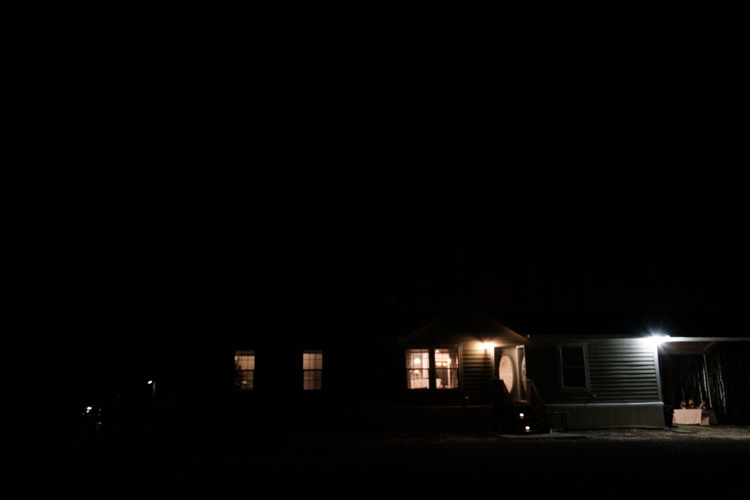 Dark nighttime exterior of a rural house with warm glowing windows and outdoor lighting.