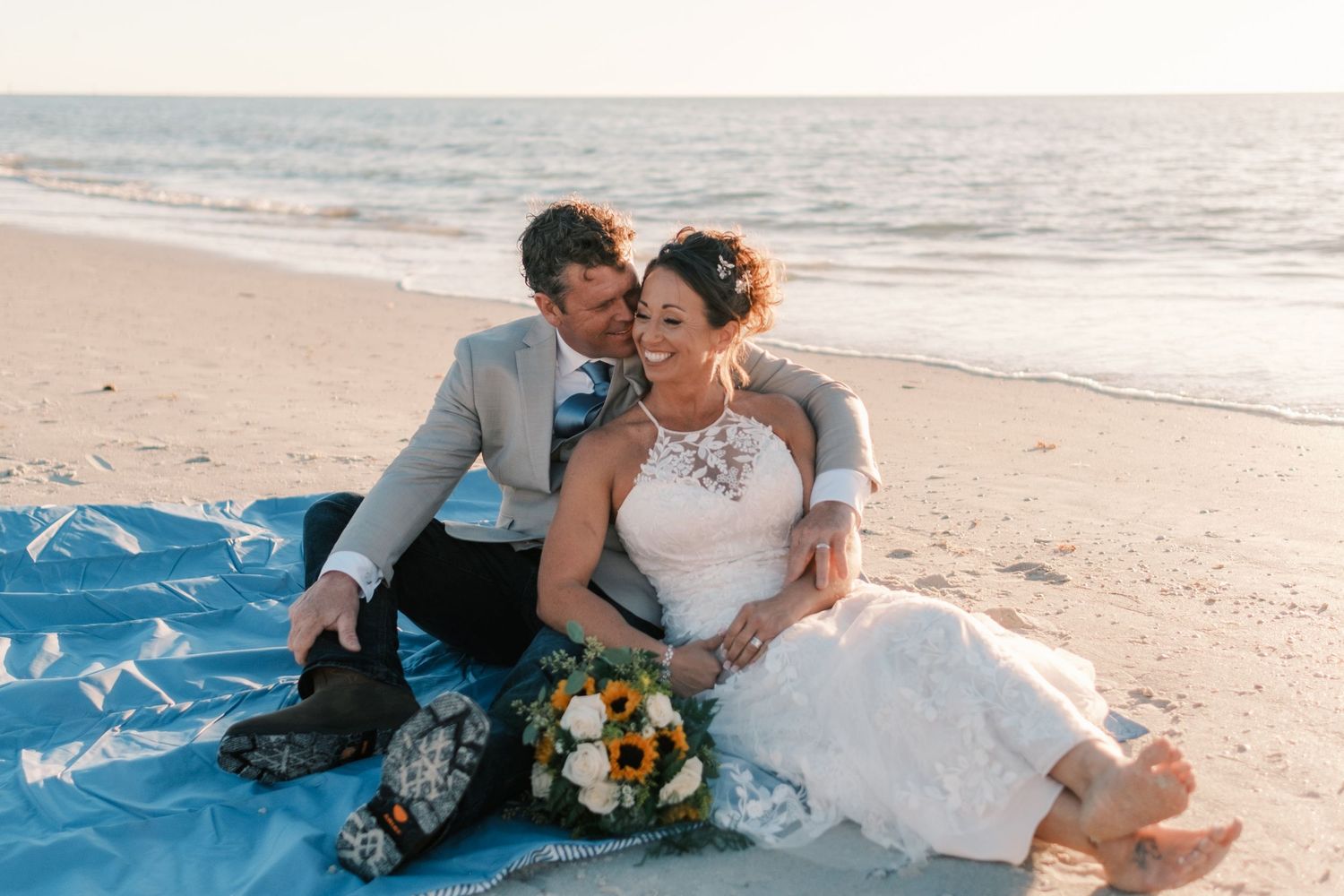 Bride and groom sitting on blue blanket on beach, sharing romantic moment at sunset with bridal bouquet.