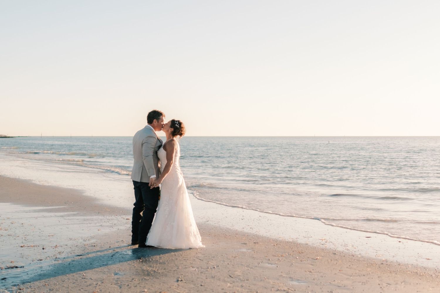 Bride and groom share a kiss at the ocean's edge during golden hour.