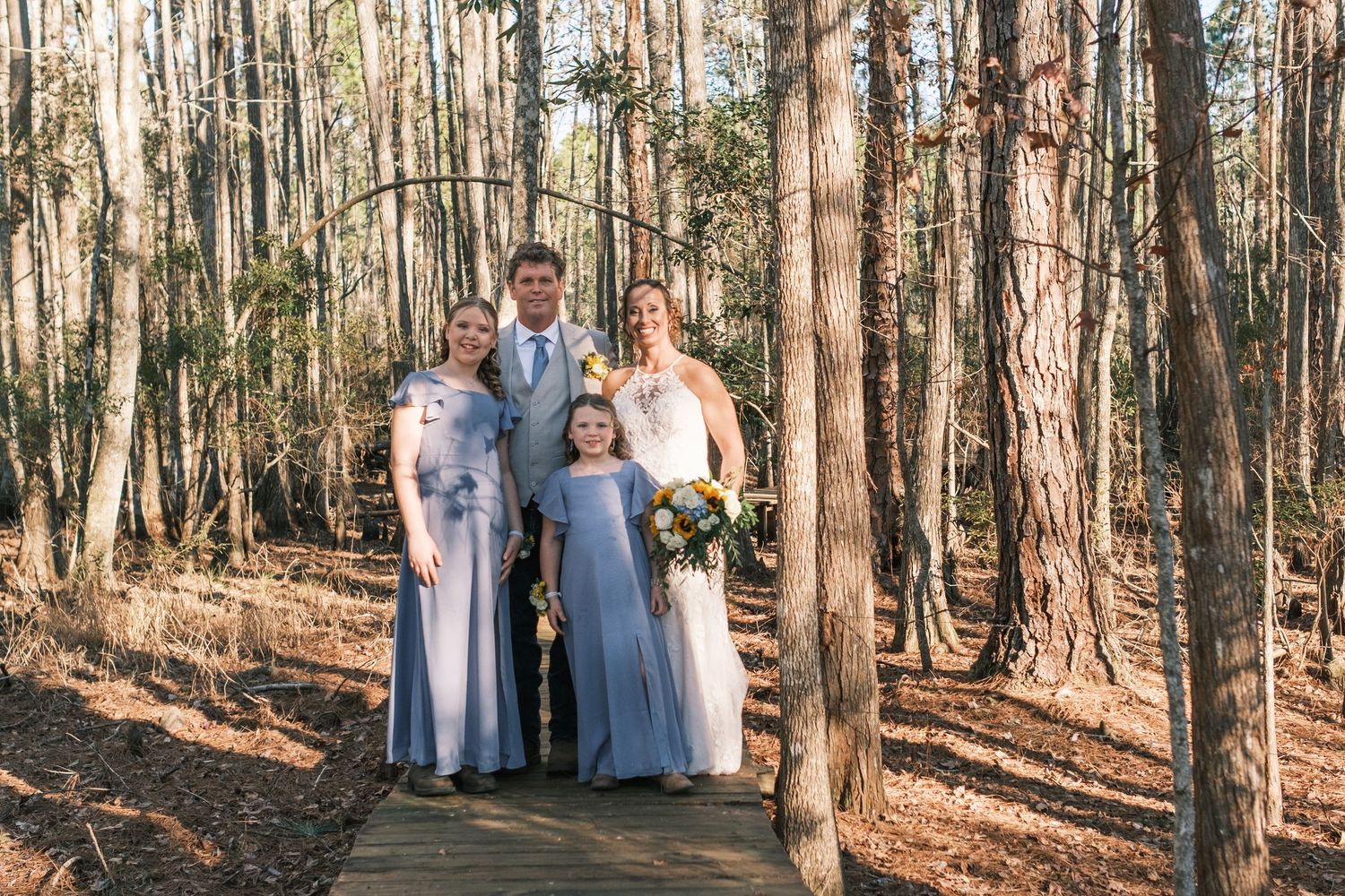 Wedding party poses on a wooden boardwalk in a pine forest, including bridesmaids and flower girl in blue dresses.