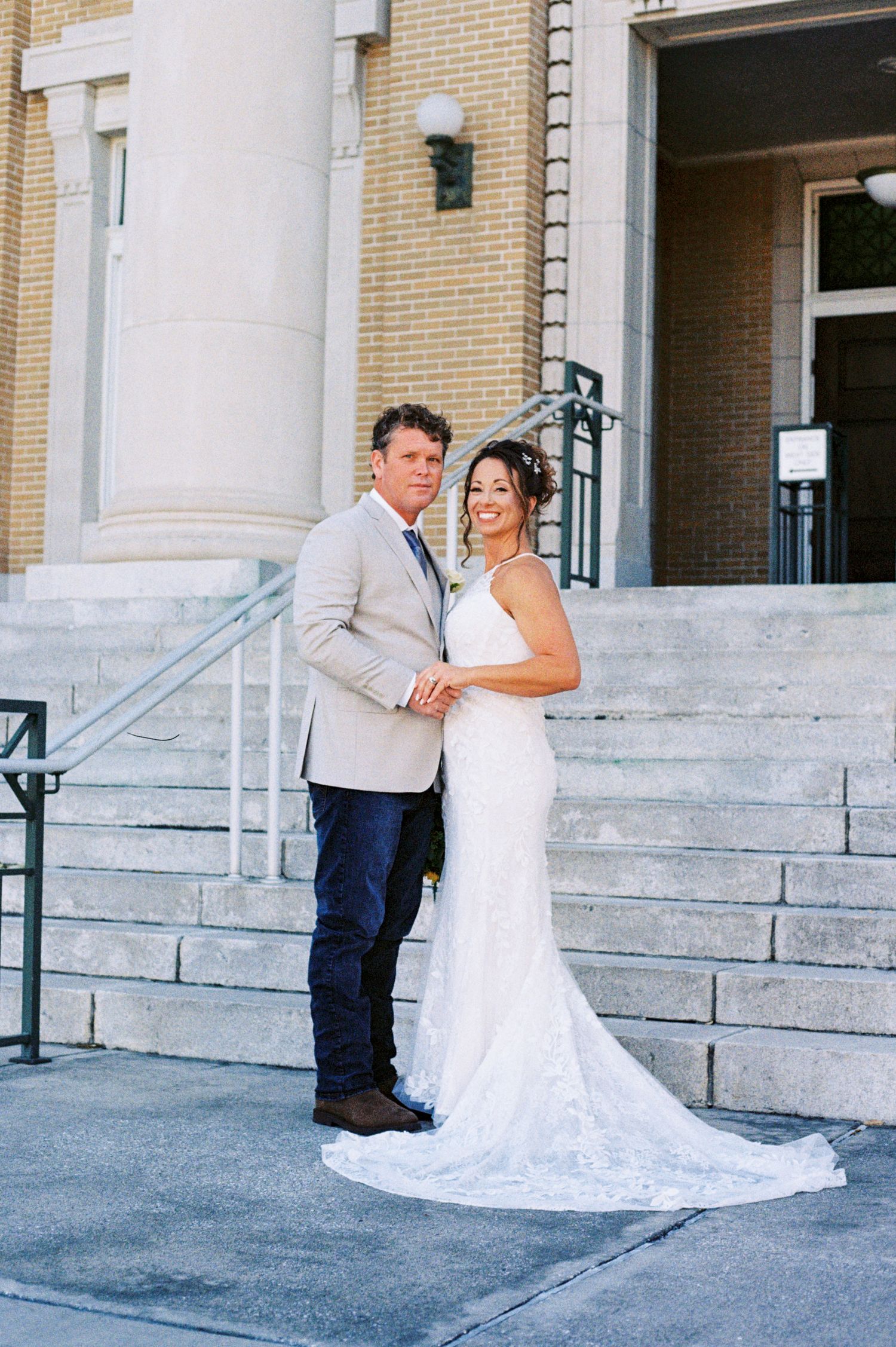 Bride and groom pose holding hands on stone steps of a classic building in wedding attire.