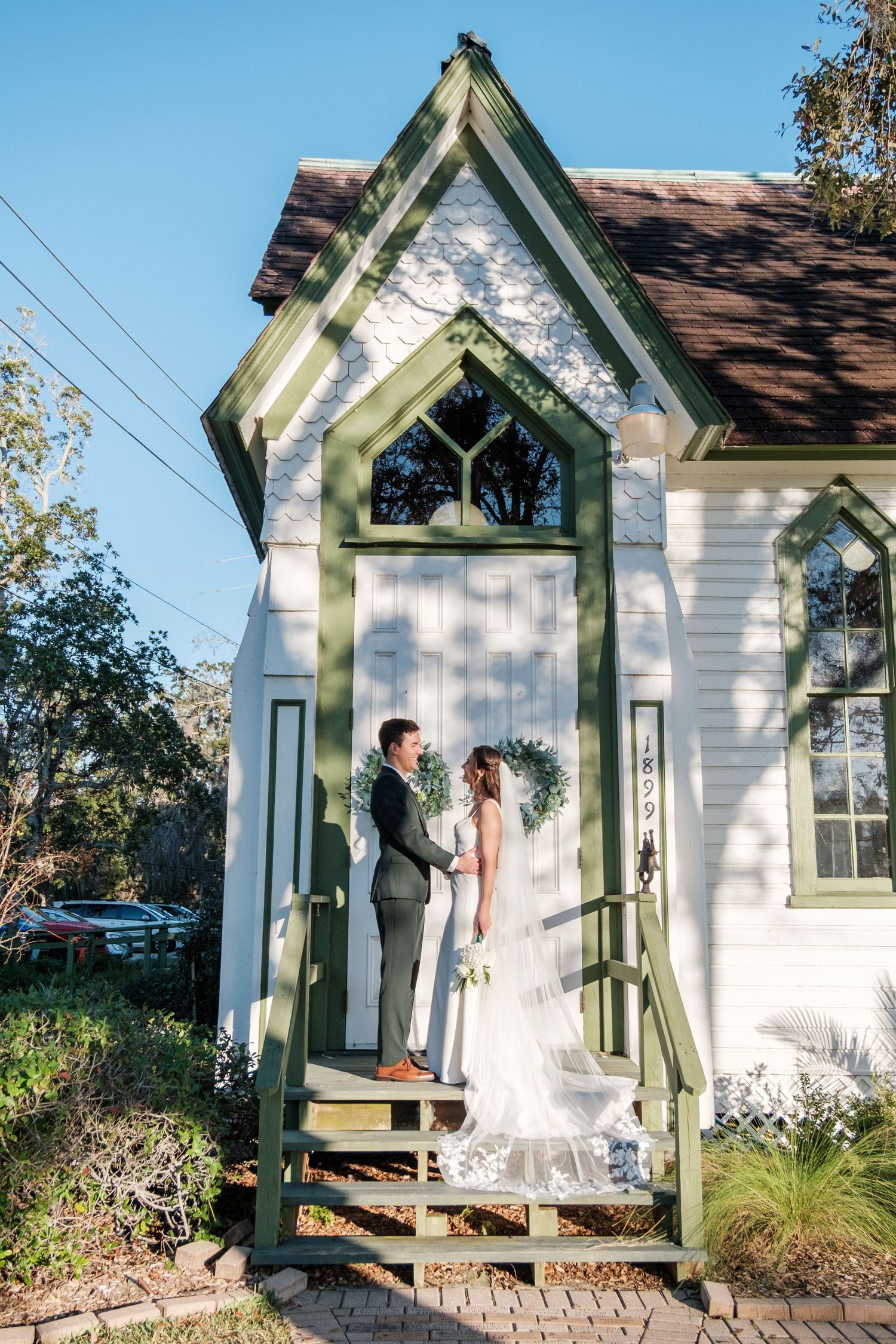A wedding couple stands in the doorway of a charming white chapel with green trim.