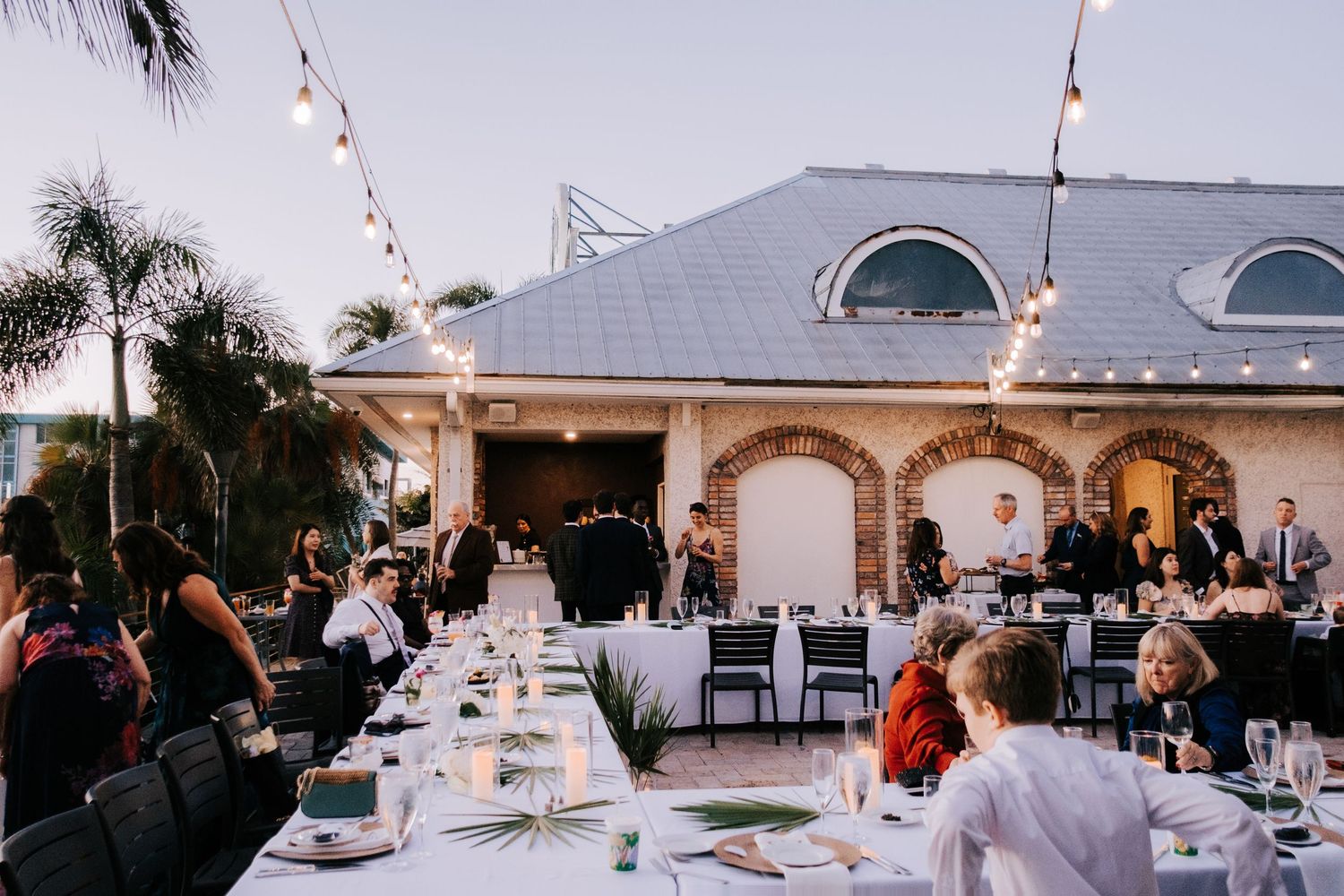 An outdoor wedding reception setup with white tents and tables decorated with string lights at dusk.