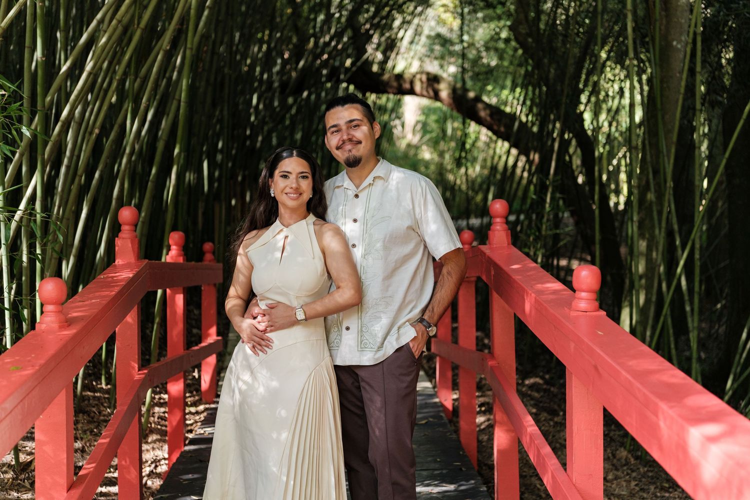 Couple posing on a red bridge surrounded by lush bamboo garden.