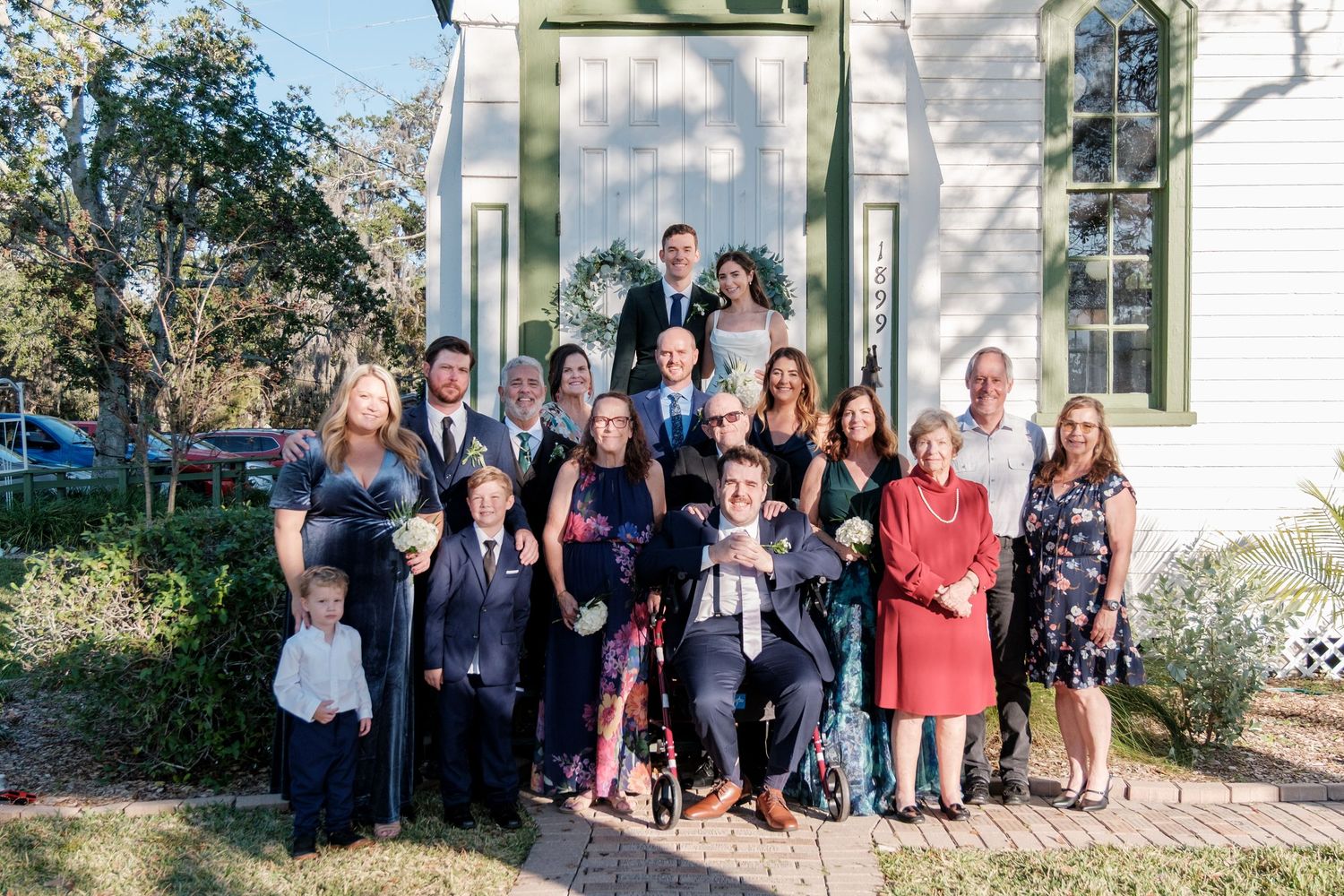 Wedding party group photo outside a white chapel with green trim.