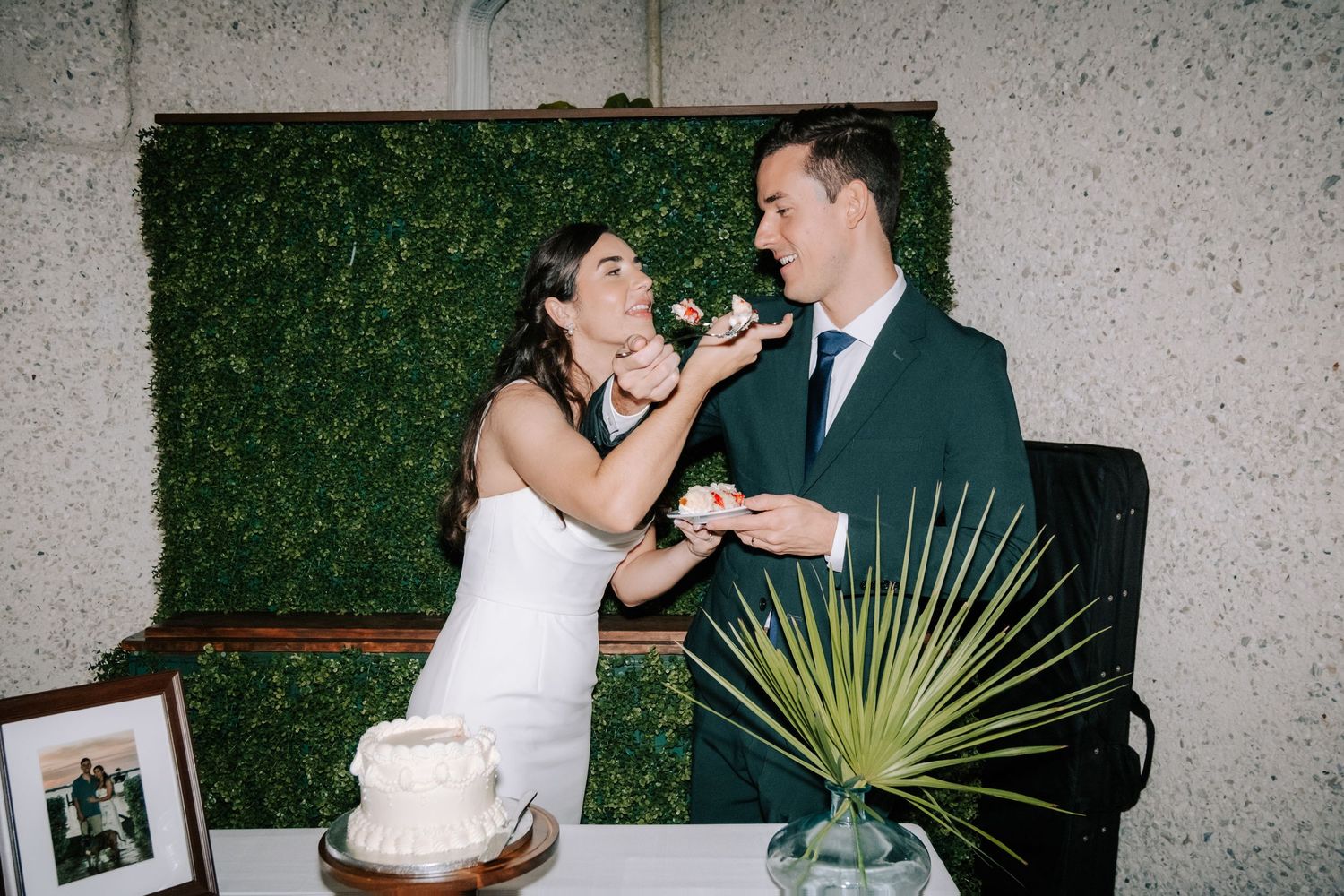 Couple sharing cake during wedding reception against green hedge backdrop.