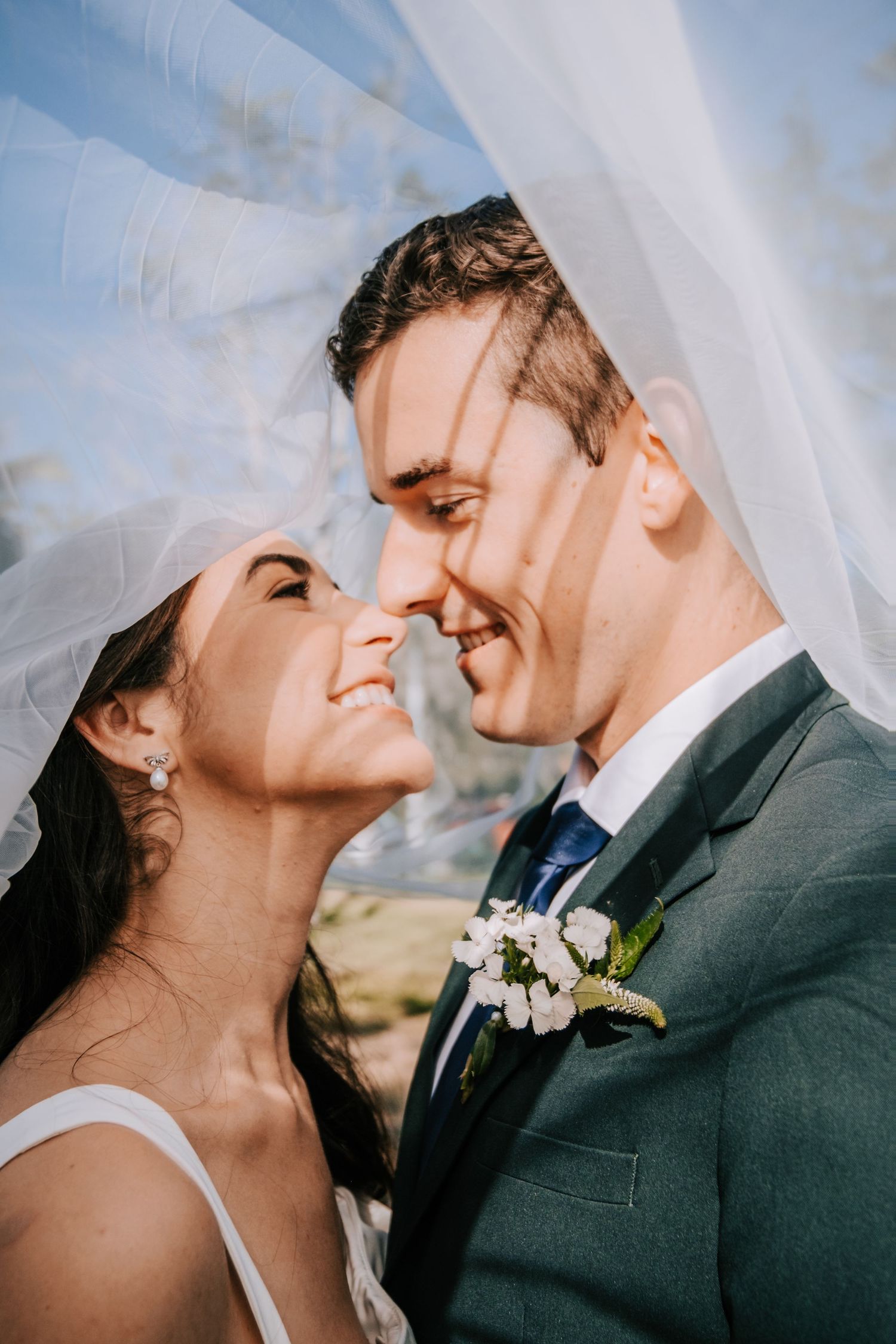 A joyful wedding portrait under flowing white veil with sunlight streaming through.