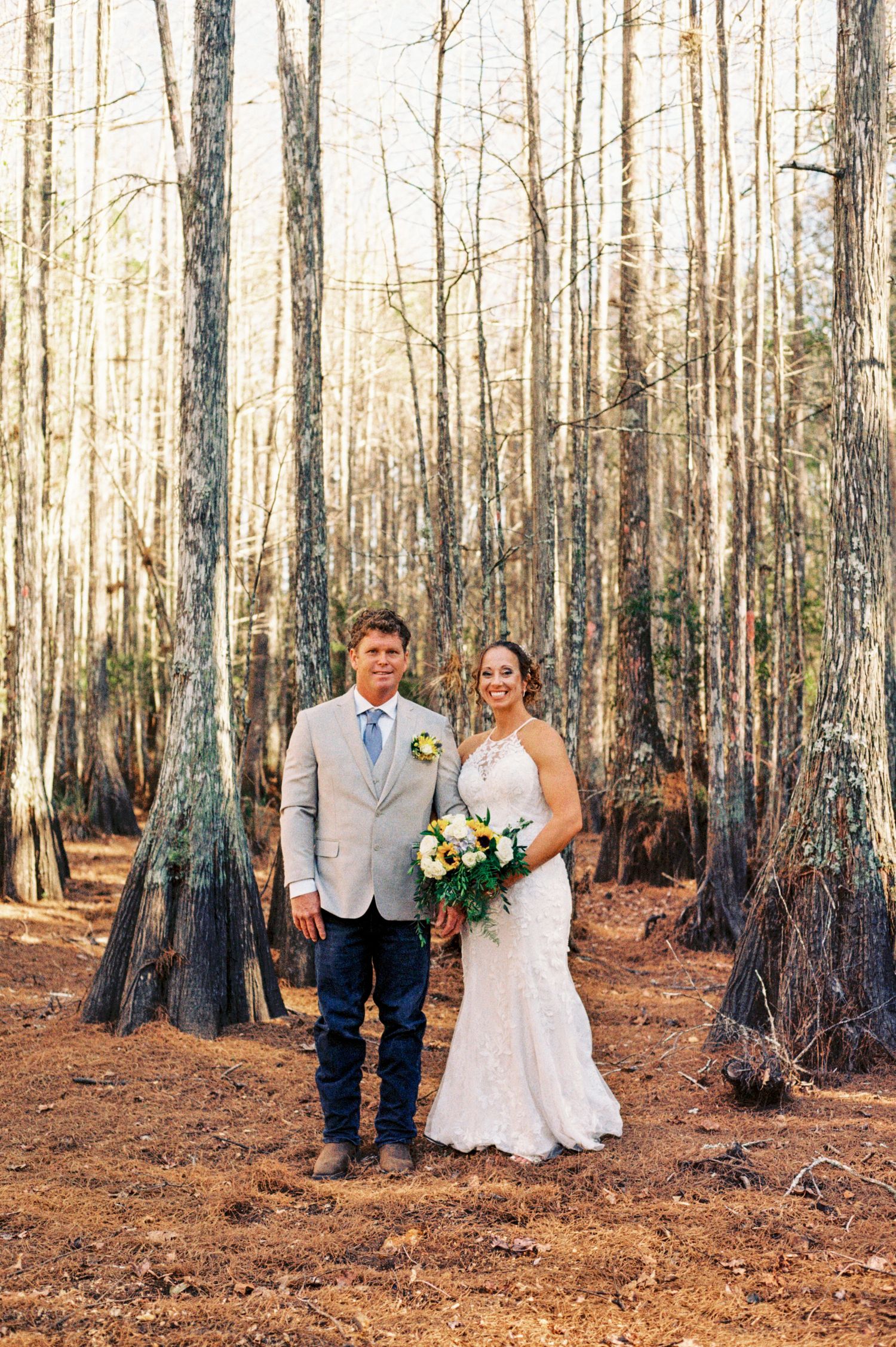 Bride in white lace gown and groom in grey suit pose together in a sunlit forest of tall bare trees.