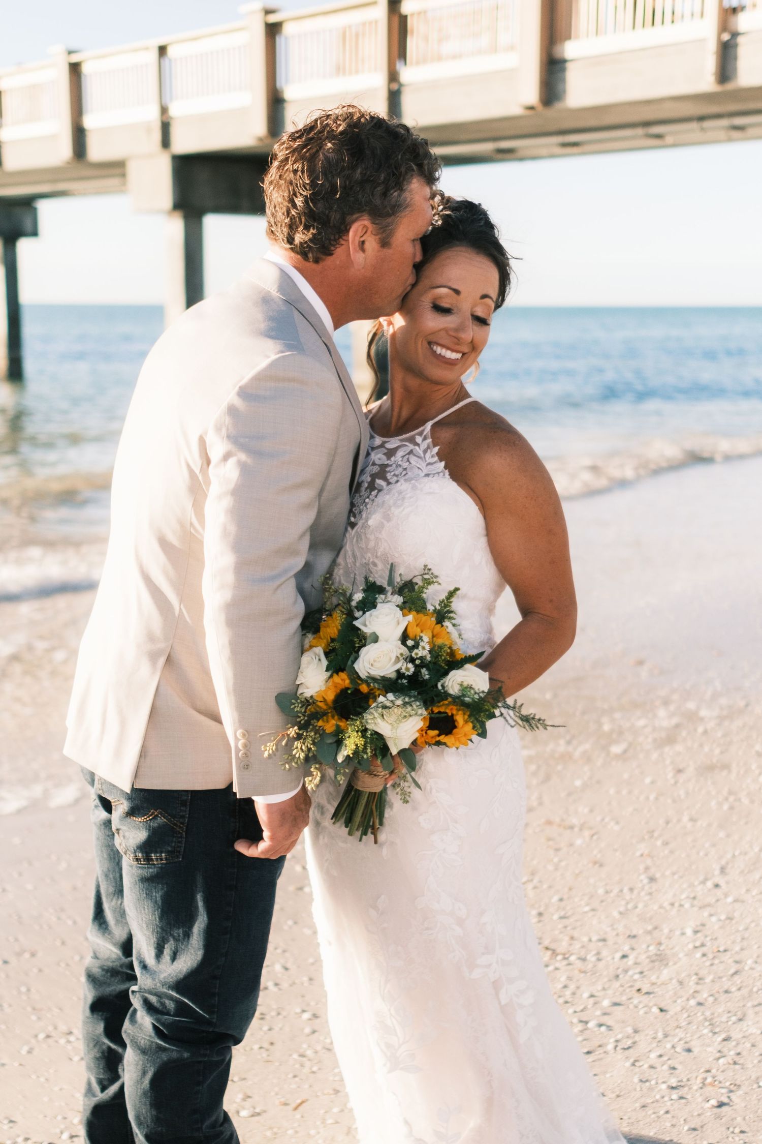 Groom kissing bride on cheek as she laughs on sunny beach.